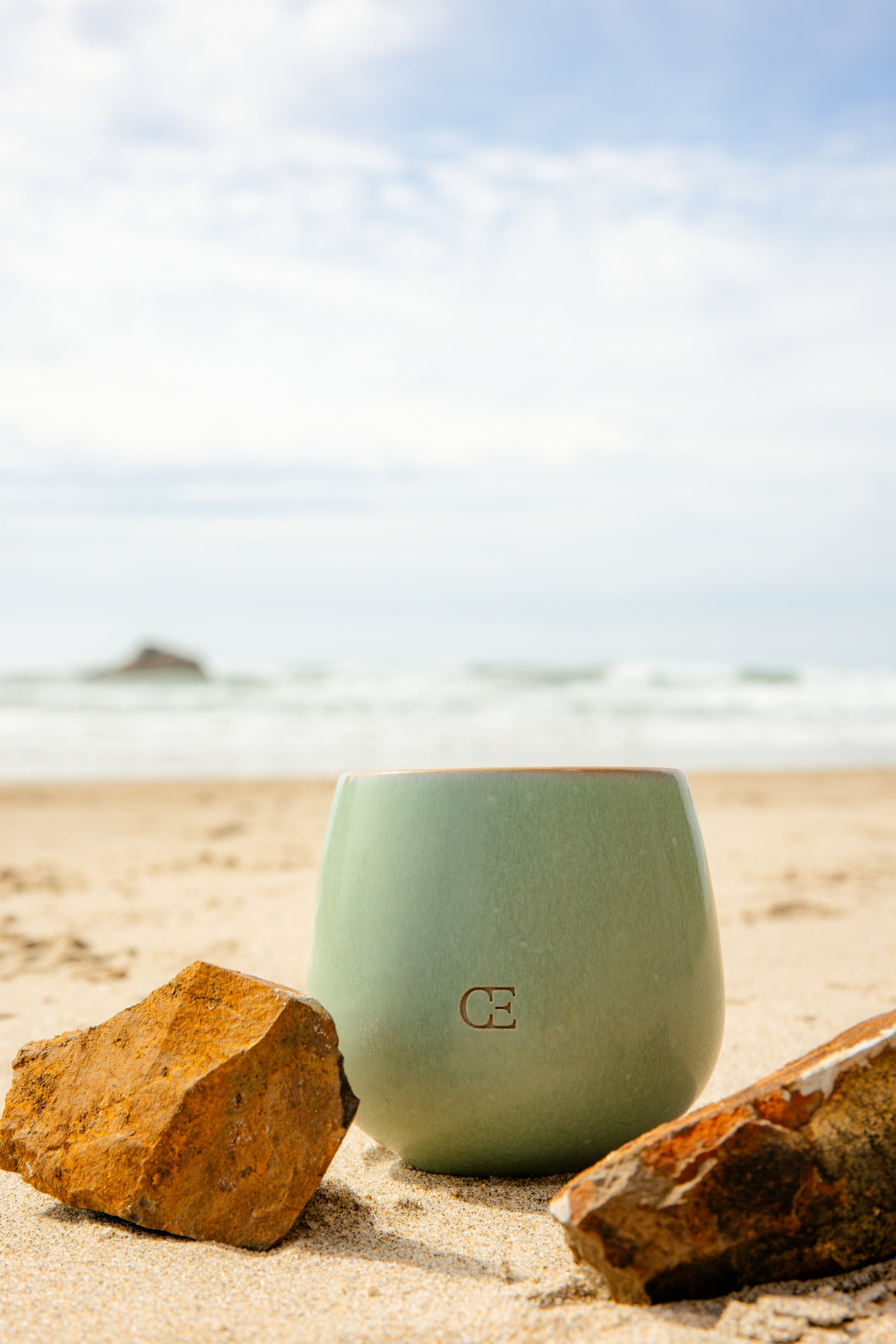 Cozy Earth Unwind Candle sits in the sand near the ocean