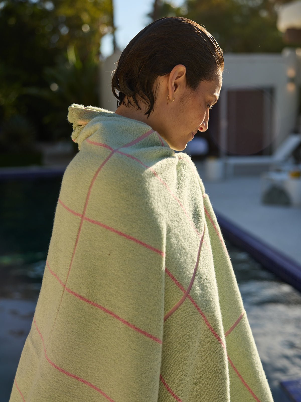 A person with short wet hair wrapped in a Cozy Earth Windowpane Resort Towel stands poolside, looking down. Sunlight filters through trees, casting warmth, with poolside furniture visible in the background. 