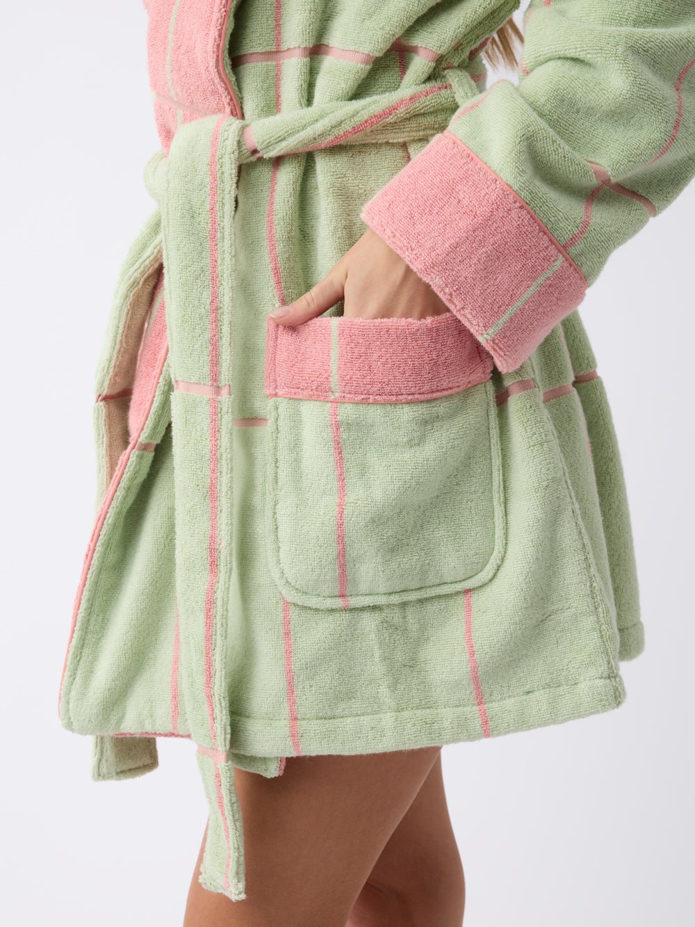 A person is wearing the Windowpane Resort Robe by Cozy Earth in pastel green with pink stripes and trim, placing a hand in the front pocket. The robe looks soft and plush. 