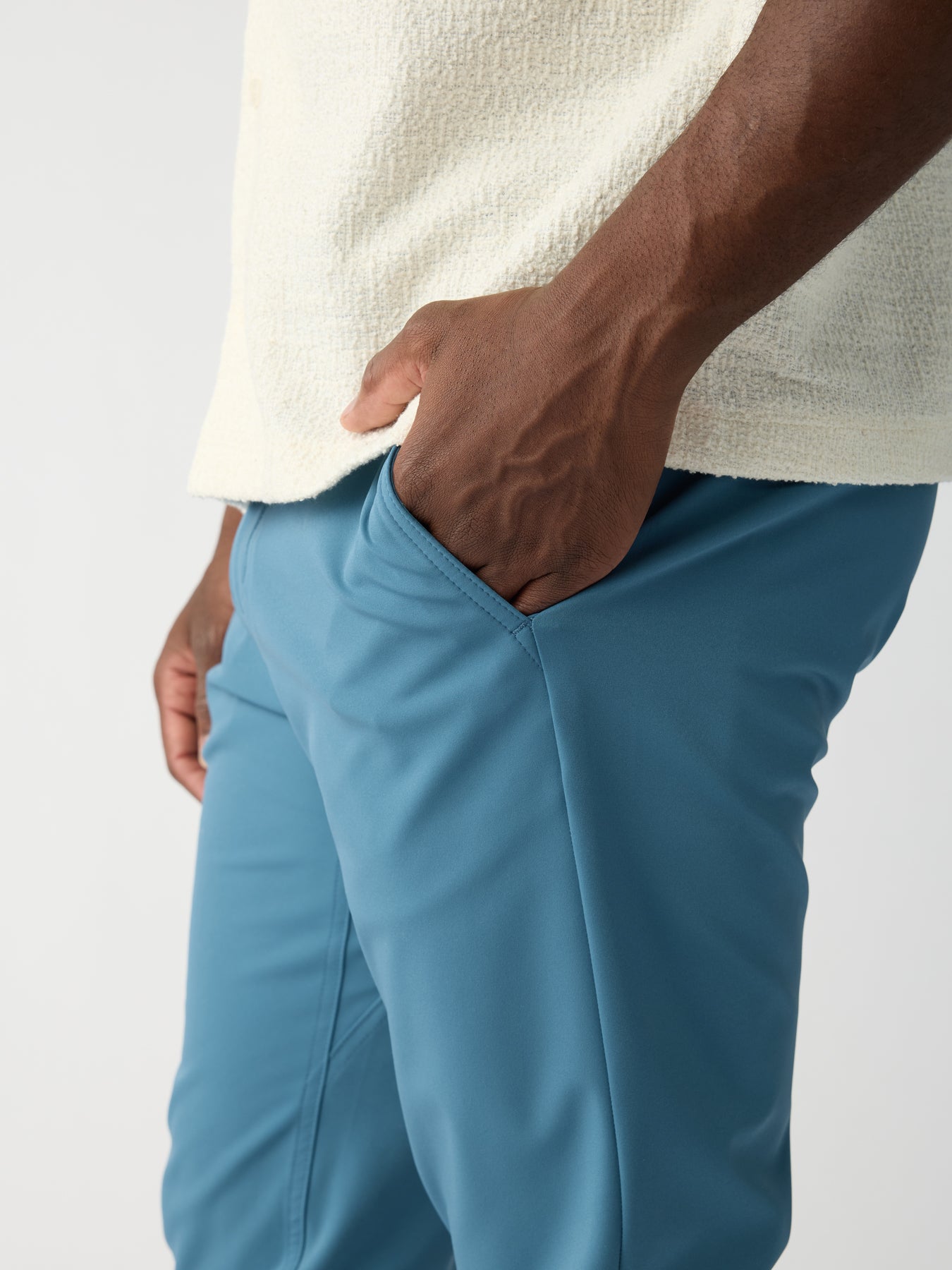 A person dressed in Cozy Earth's Men's Everywhere Pant, featuring blue pants and a textured white top, stands against a plain background with one hand in the pocket, highlighting the clothing details and a relaxed pose. 