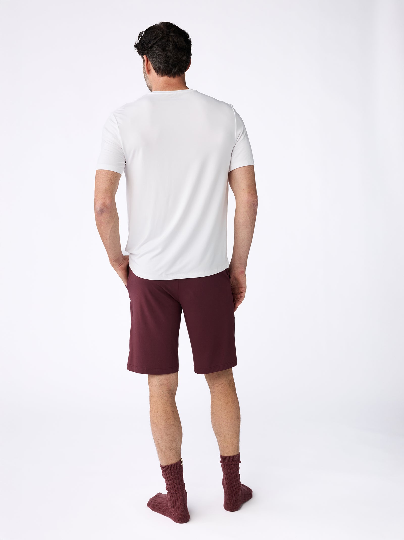 A man stands facing away, wearing a Men's Stretch-Knit Bamboo Lounge Tee from Cozy Earth in white, along with burgundy shorts and matching burgundy socks. The plain white background highlights the relaxed ensemble. 