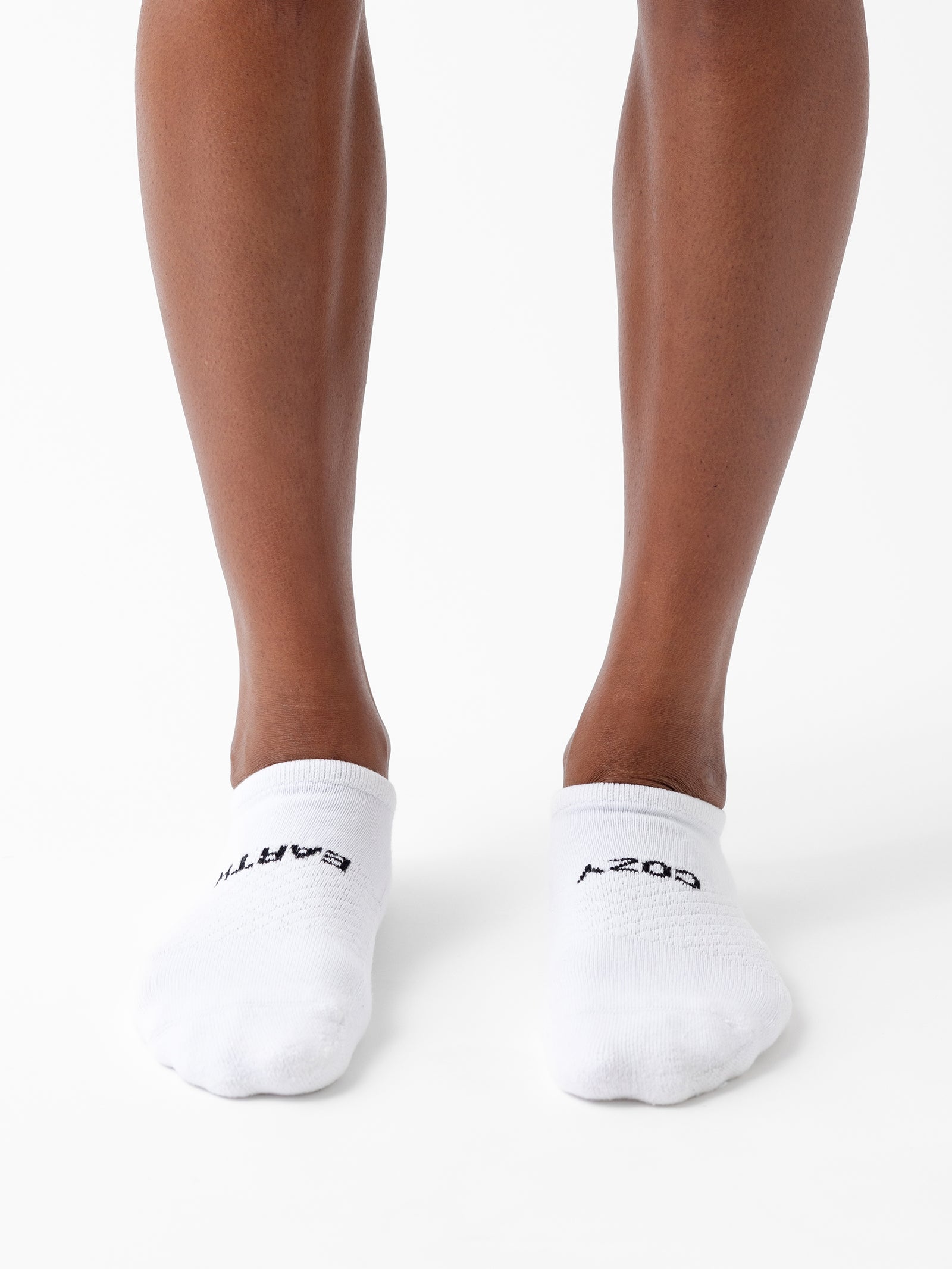 A person wearing Cozy Earth's Essential Ankle Sock 2-Pack is standing on a white surface. The white socks feature black text, with "ARTA" on the left and "A-207" on the right. The person's skin is brown. 