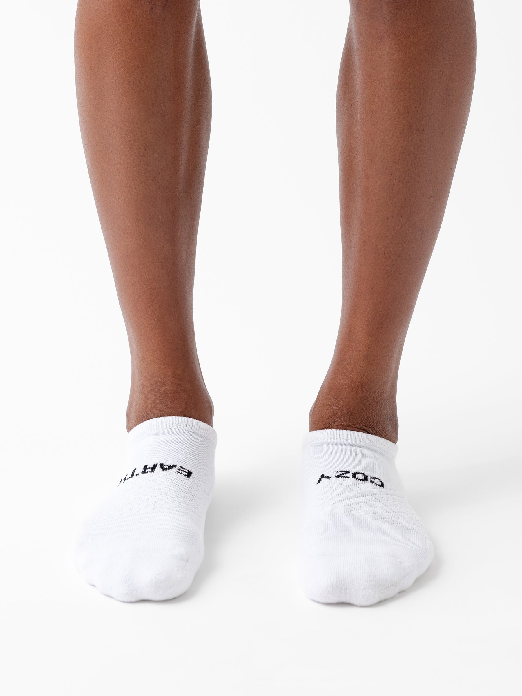 A person stands on a white surface wearing Cozy Earth Essential Ankle Sock 8-Pack in white with black "BARTY" and "A.204" text. Only their lower legs and feet are visible. 