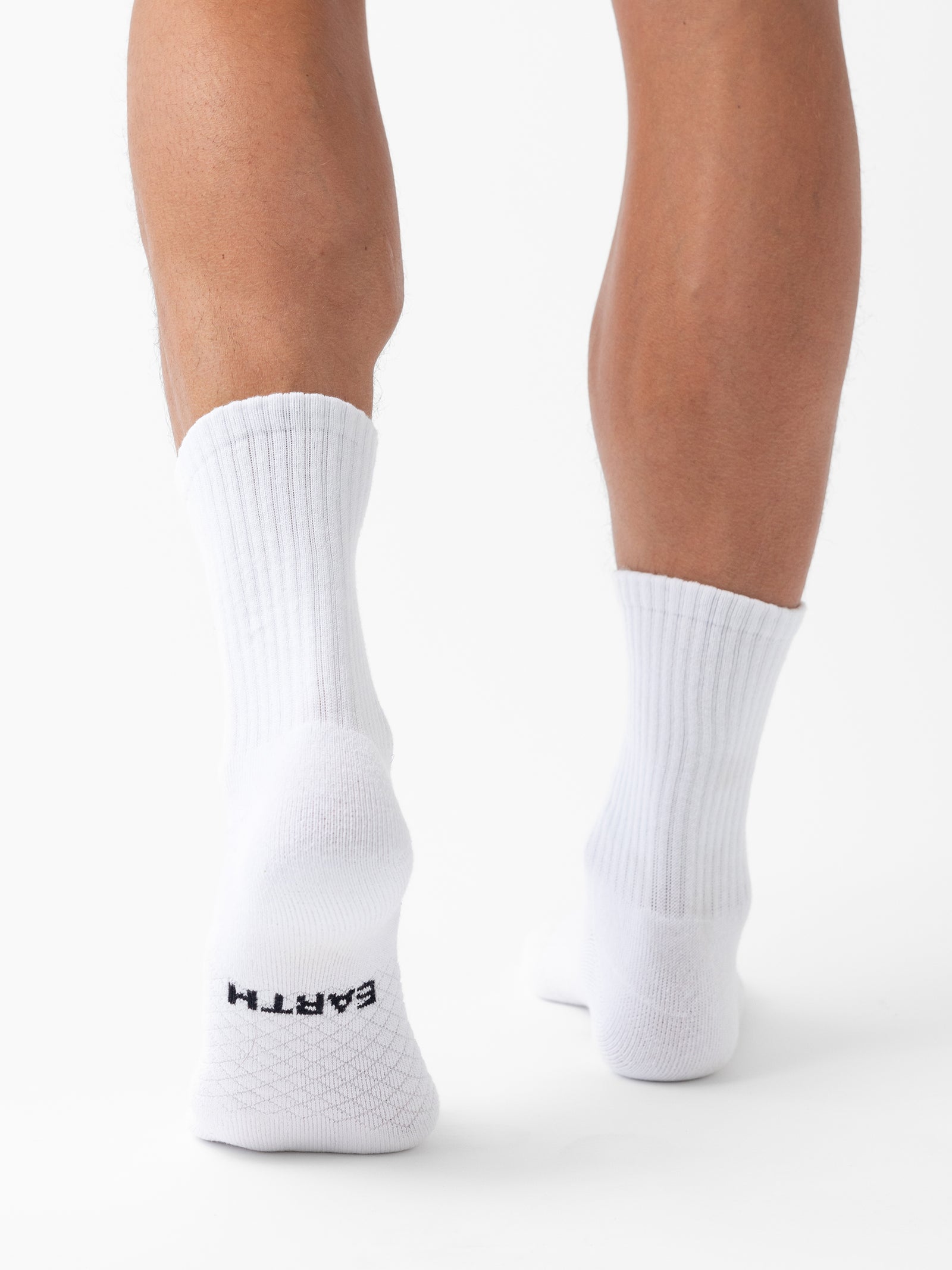 A person wearing white Essential Calf Socks from the Cozy Earth 4-Pack stands on a white background, facing away from the camera. The left sock has the word "EARTH" written in black letters near the bottom. The background is plain and evenly lit. 