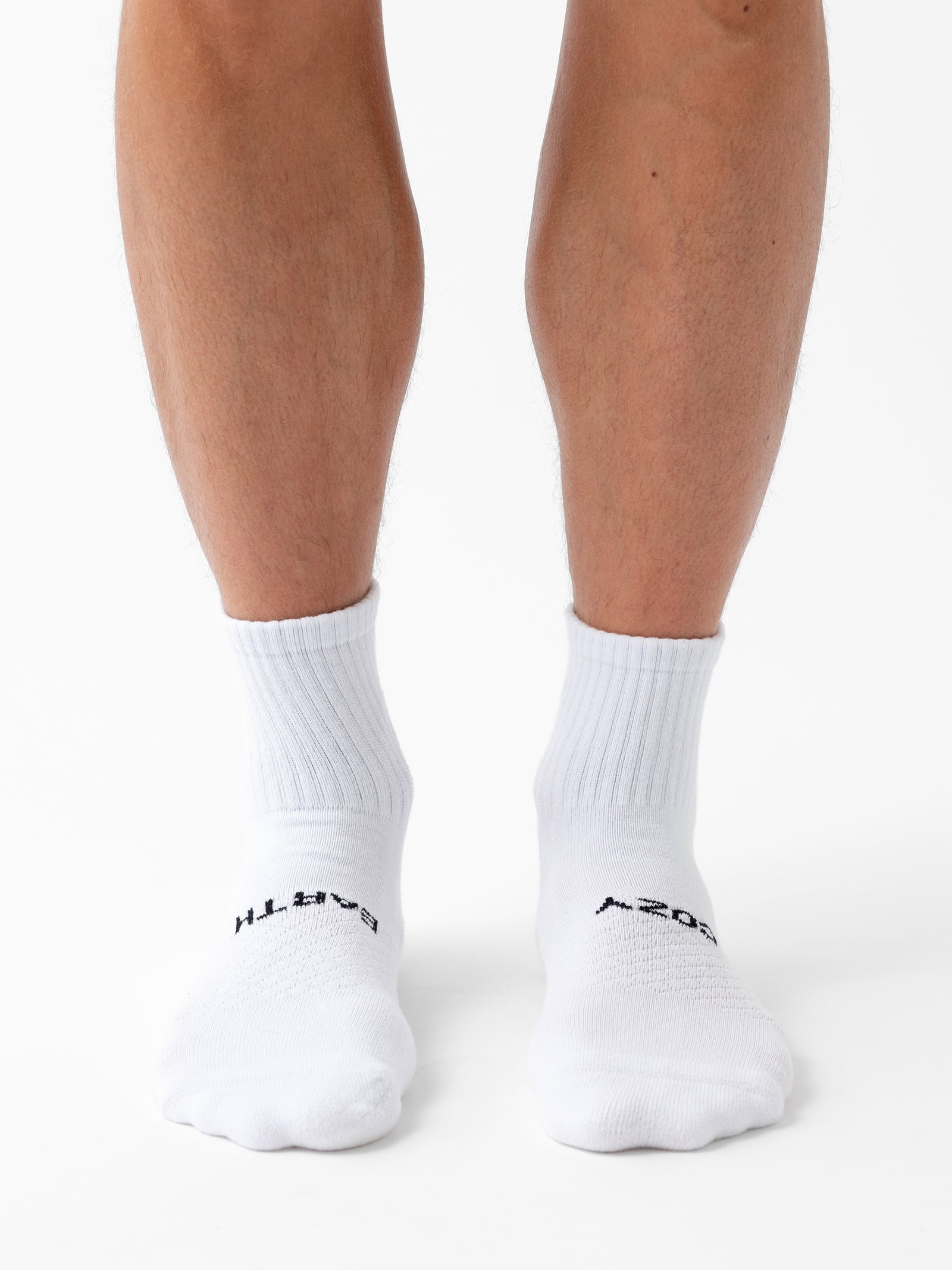 A person is standing with legs slightly apart, wearing Cozy Earth's Essential Quarter Sock from their 2-Pack. The white mid-calf socks feature black text near the toes against a plain white background. 