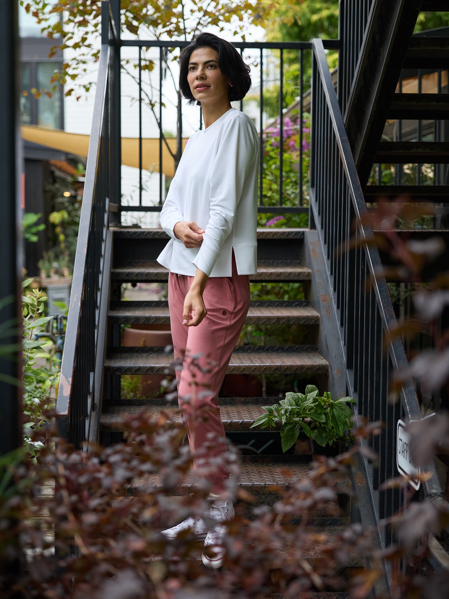 A person with short dark hair stands on a metal staircase amidst lush greenery. They are wearing a white Cozy Earth Women's StudioLite Crewneck and pink pants, with one hand in their pocket. The setting appears to be an outdoor garden or courtyard area with plants and trees around. 