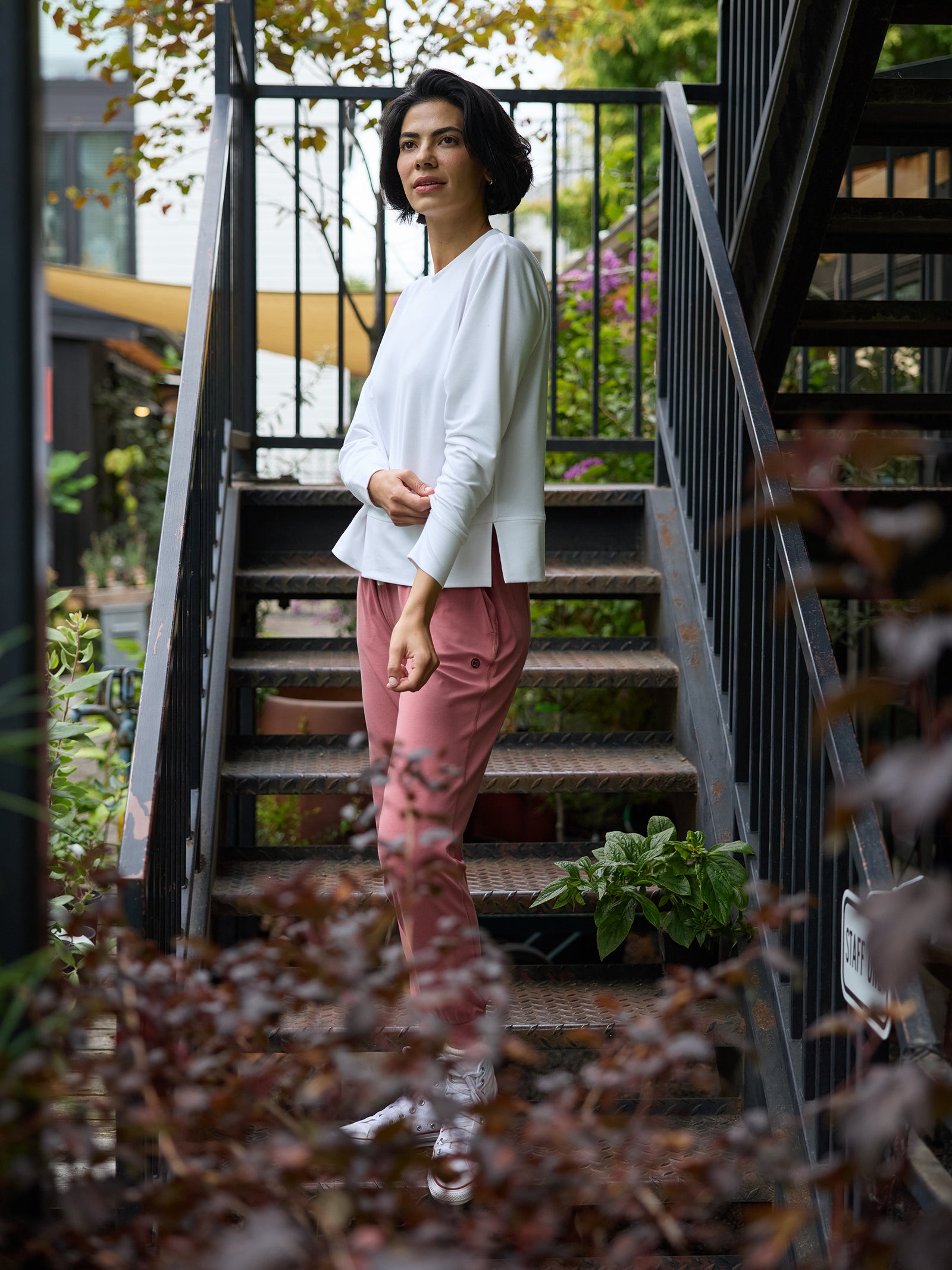 A person with short dark hair stands on a metal staircase amidst lush greenery. They are wearing a white Cozy Earth Women's StudioLite Crewneck and pink pants, with one hand in their pocket. The setting appears to be an outdoor garden or courtyard area with plants and trees around. 