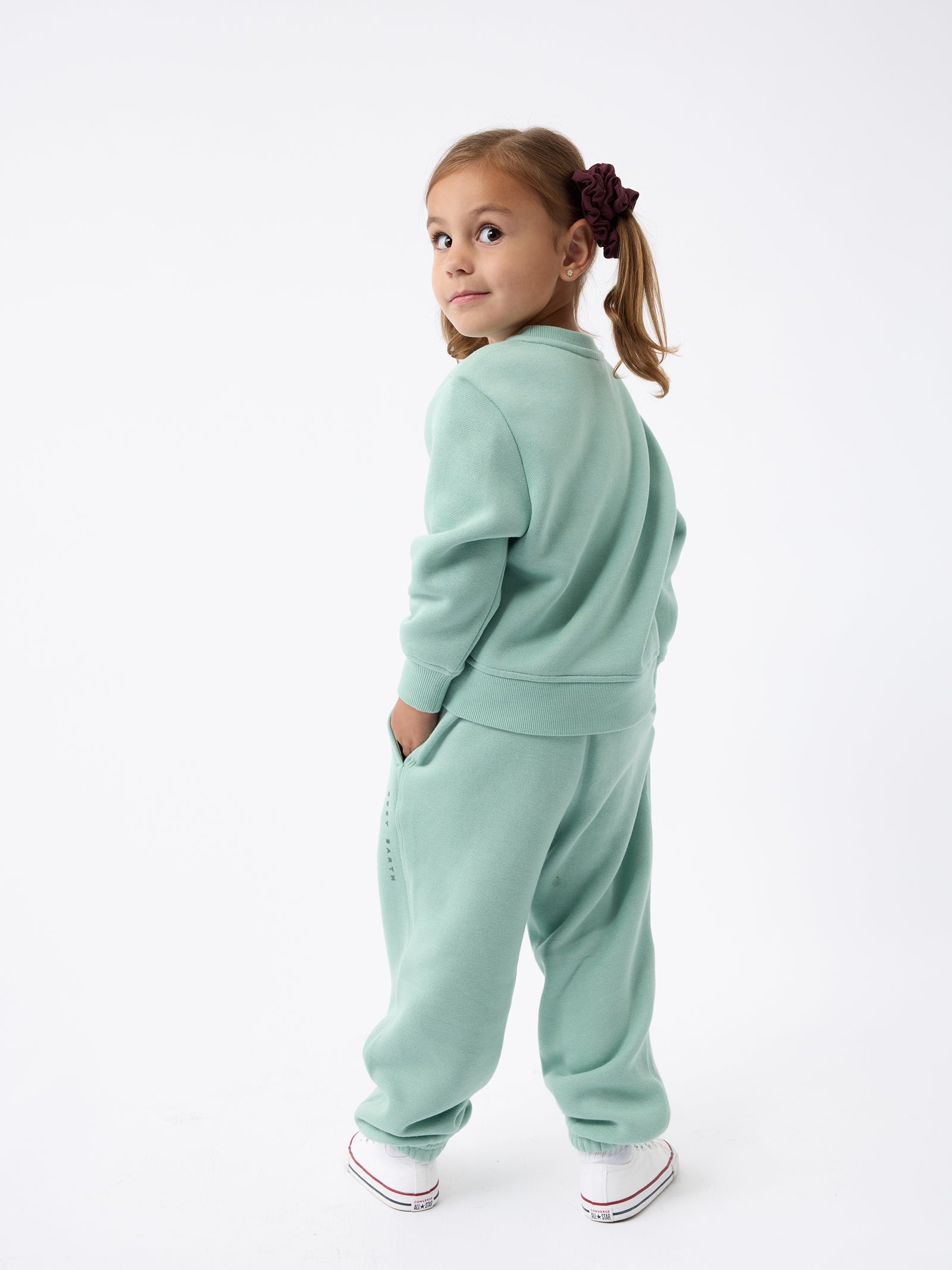 A young girl stands facing away, glancing back with a smile. She is dressed in a Kid's CityScape Crewneck by Cozy Earth paired with white sneakers, and her hair is styled in two pigtails secured with hair ties. The backdrop is white and minimalistic. 