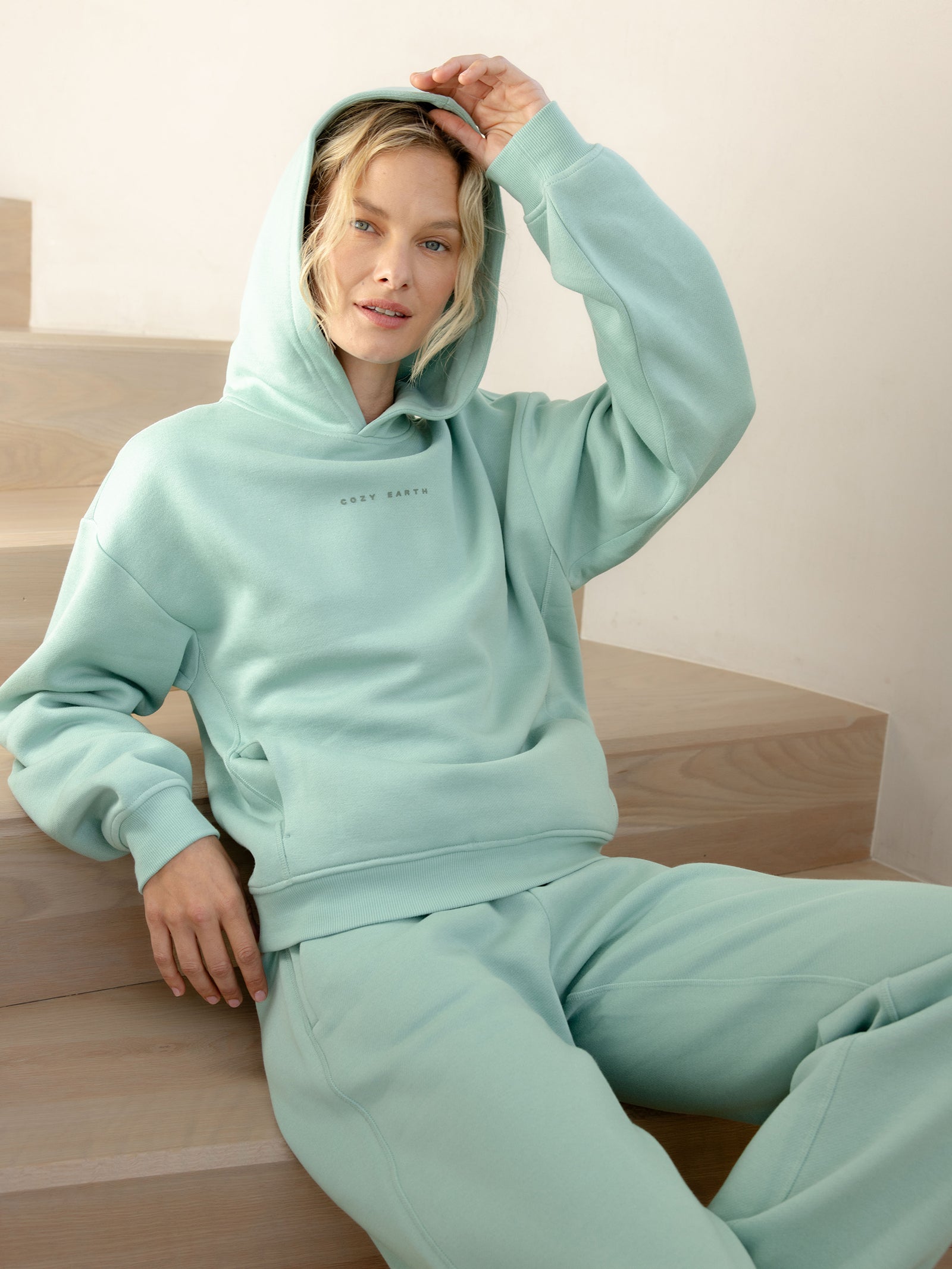 A person sits on wooden stairs, dressed in a mint green Women's CityScape Hoodie and matching sweatpants by Cozy Earth. They have light hair and are holding the hood of the hoodie with one hand, looking directly at the camera in a minimal and softly lit setting. 