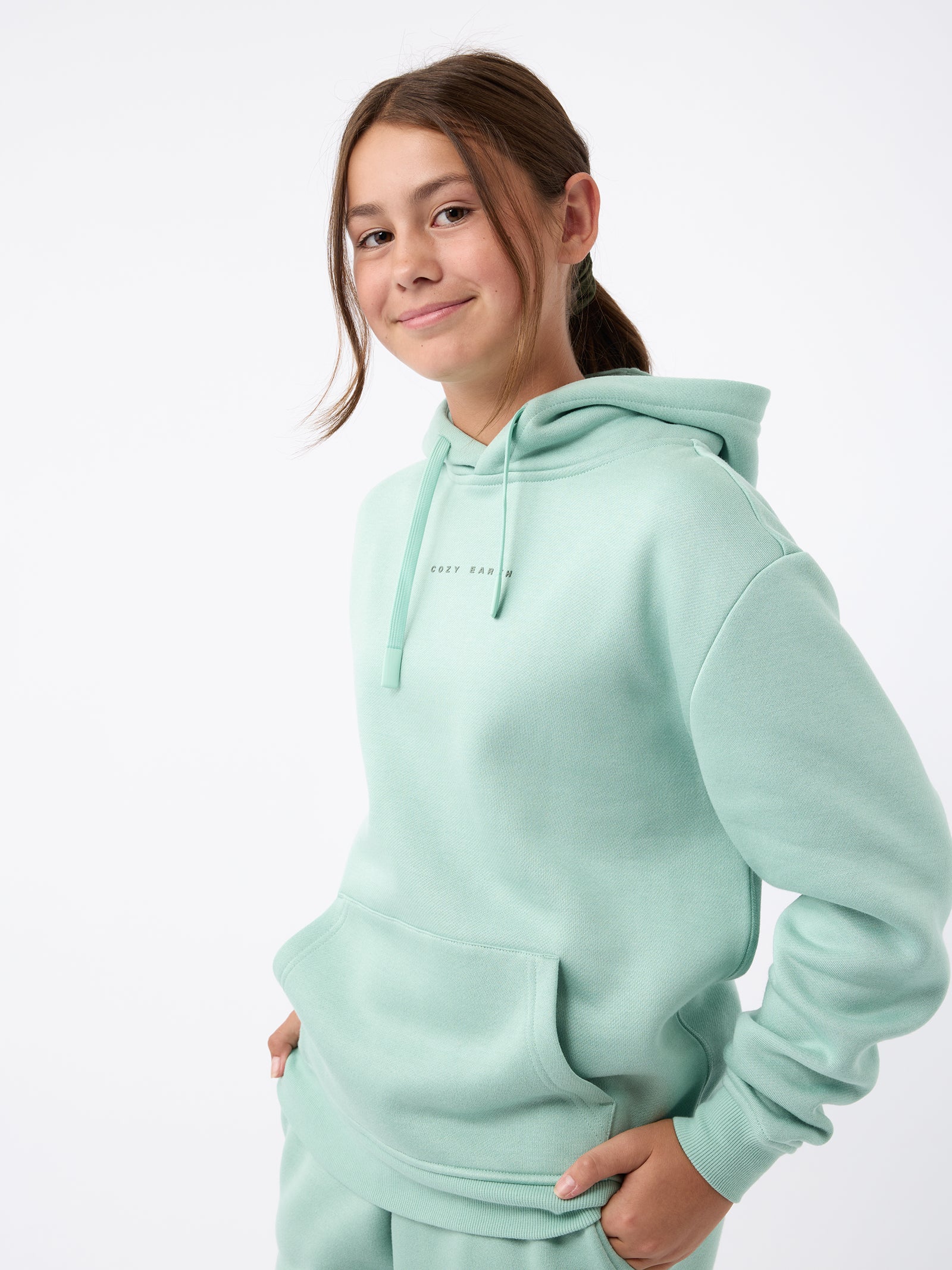 A young person wearing Cozy Earth's Kid's CityScape Hoodie in mint green smiles while standing against a plain white background. The hoodie features a front pocket and a subtle text design. They have one hand in the pocket and casually loose hair. 