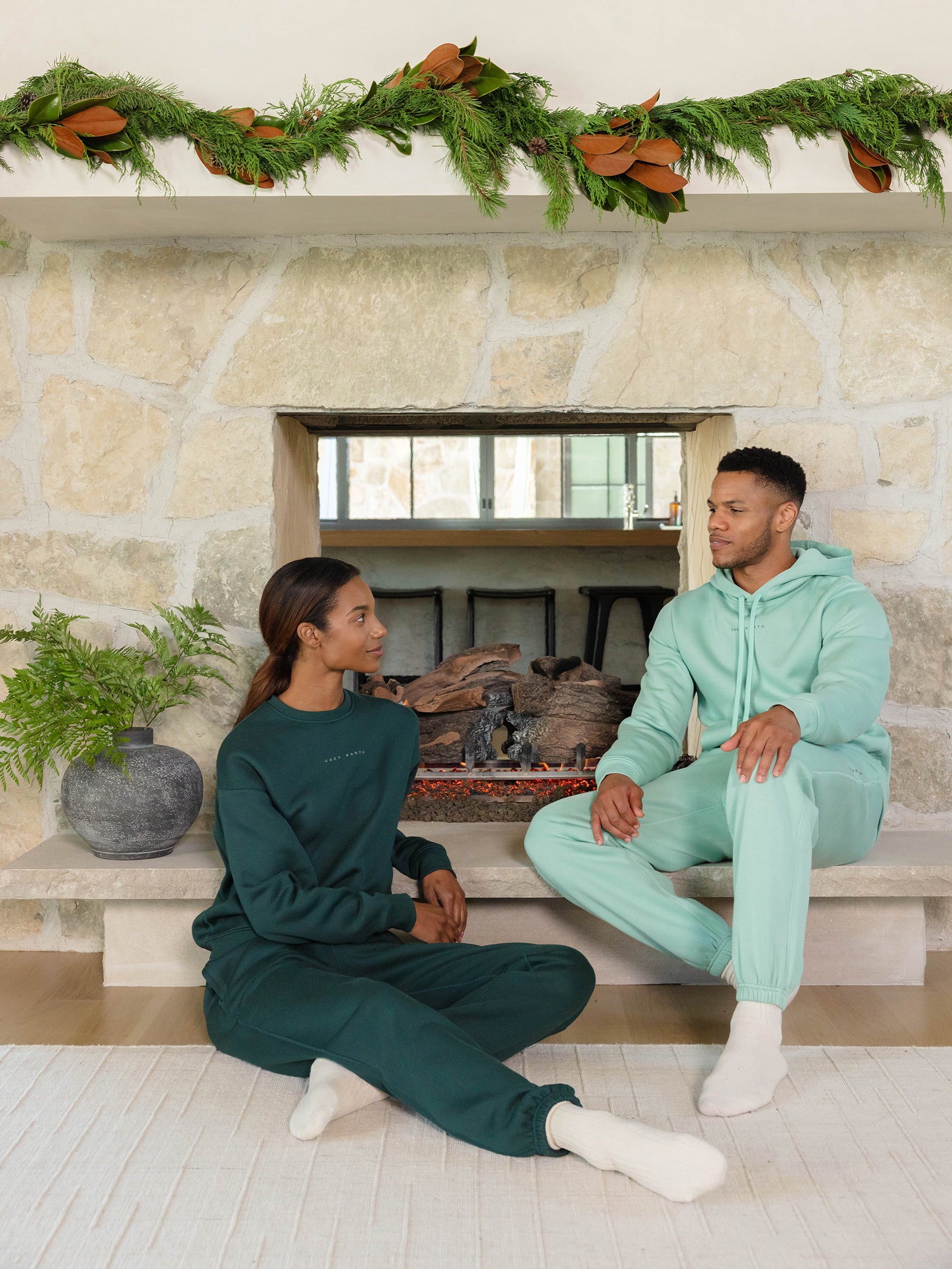 A man and woman dressed in matching green CityScape Hoodies from Cozy Earth sit together in front of a stone fireplace. Above the fireplace, a garland is draped, and a potted plant is placed nearby. They gaze at each other, enveloped in the room's cozy atmosphere. 