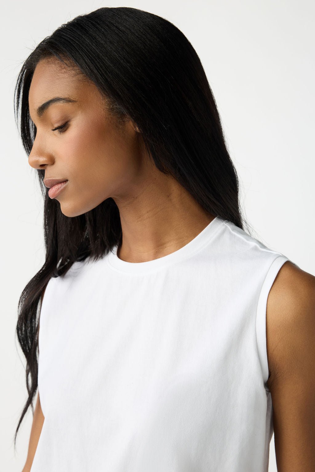 A woman with long dark hair dons a Cozy Earth Women's All Day Sleeveless Tee, appearing calm as she gazes slightly downward against a neutral background.