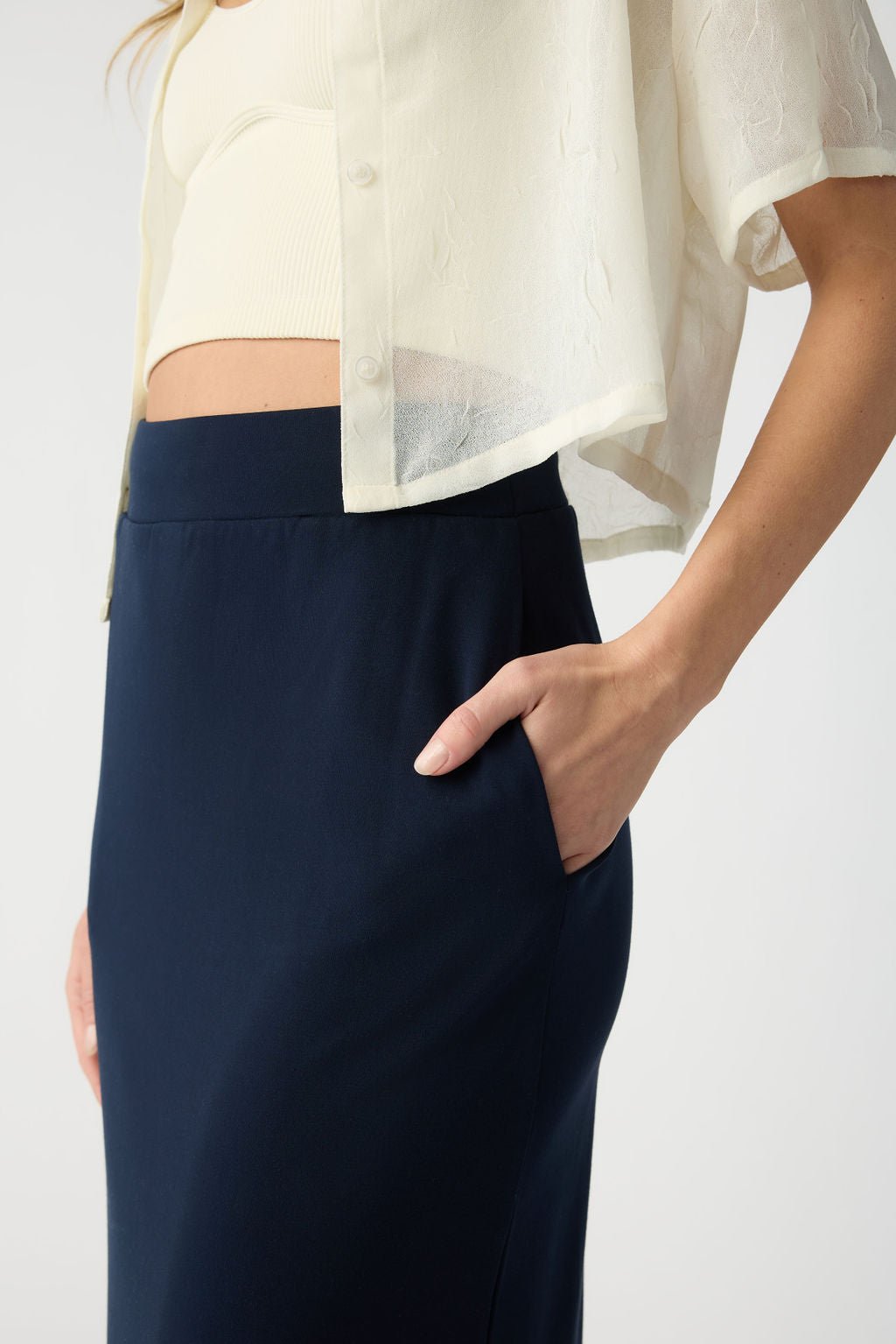 A person is shown from neck to mid-thigh wearing a white cropped blouse and cream top, with their right hand in the pocket of a dark blue Cozy Earth Women's Brushed Bamboo Midi Skirt. 