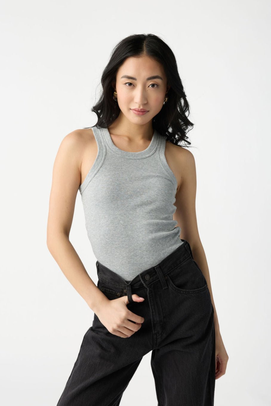 A dark haired woman wears Cozy Earth Women's Fine Ribbed Tank in Heather Grey. She holds the belt loop of her black jeans and stands in front of a white background. 