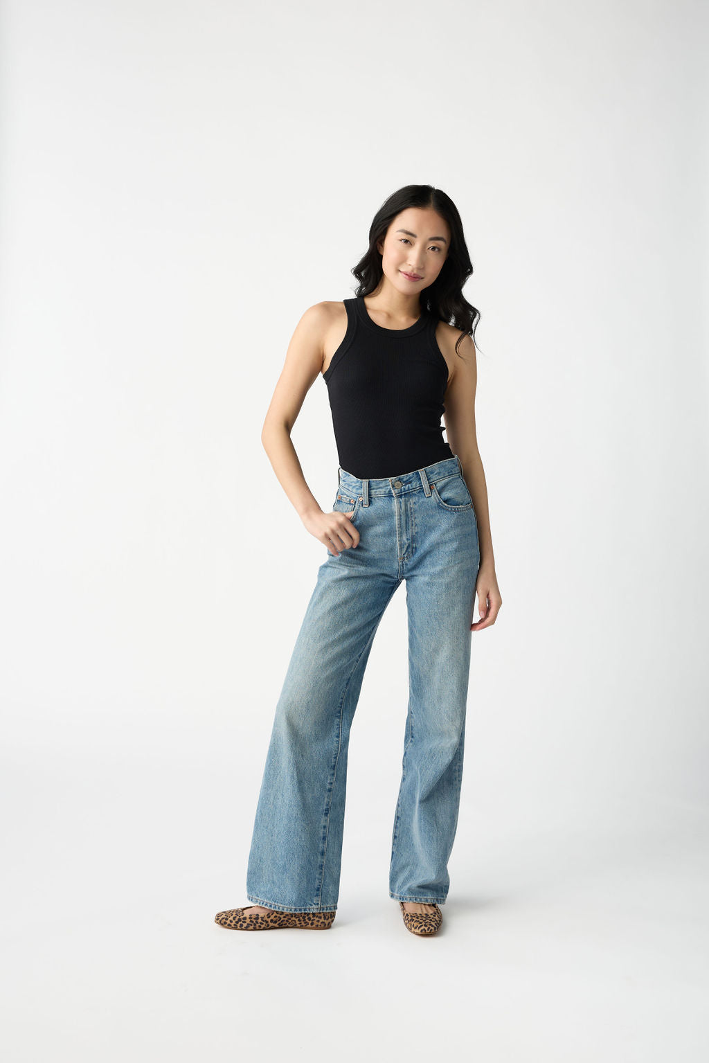 A dark haired woman wears Cozy Earth Women's Fine Ribbed Tank in Jet Black. She puts one thumb in the pocket of her blue jeans and stands in front of a white background. 