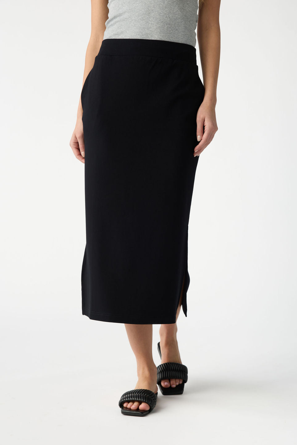 A person in a light gray top, Cozy Earth's Women's Brushed Bamboo Midi Skirt in black with a side slit, and black woven slide sandals stands against a plain white background. 