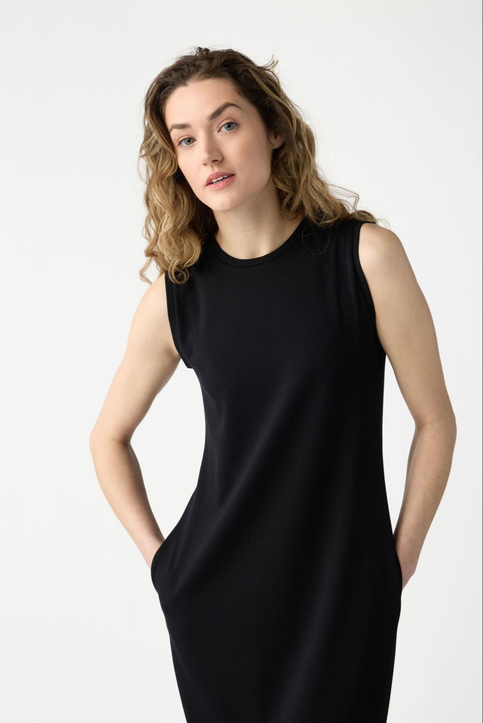 A person with long, wavy hair is wearing the Women's Brushed Bamboo Sleeveless Midi Dress by Cozy Earth in black, hands in pockets. They stand against a plain white background, looking slightly upwards with a neutral expression. 