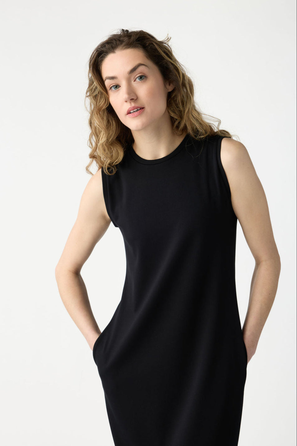 A person with long, wavy hair is wearing the Women's Brushed Bamboo Sleeveless Midi Dress by Cozy Earth in black, hands in pockets. They stand against a plain white background, looking slightly upwards with a neutral expression. |Color:Black