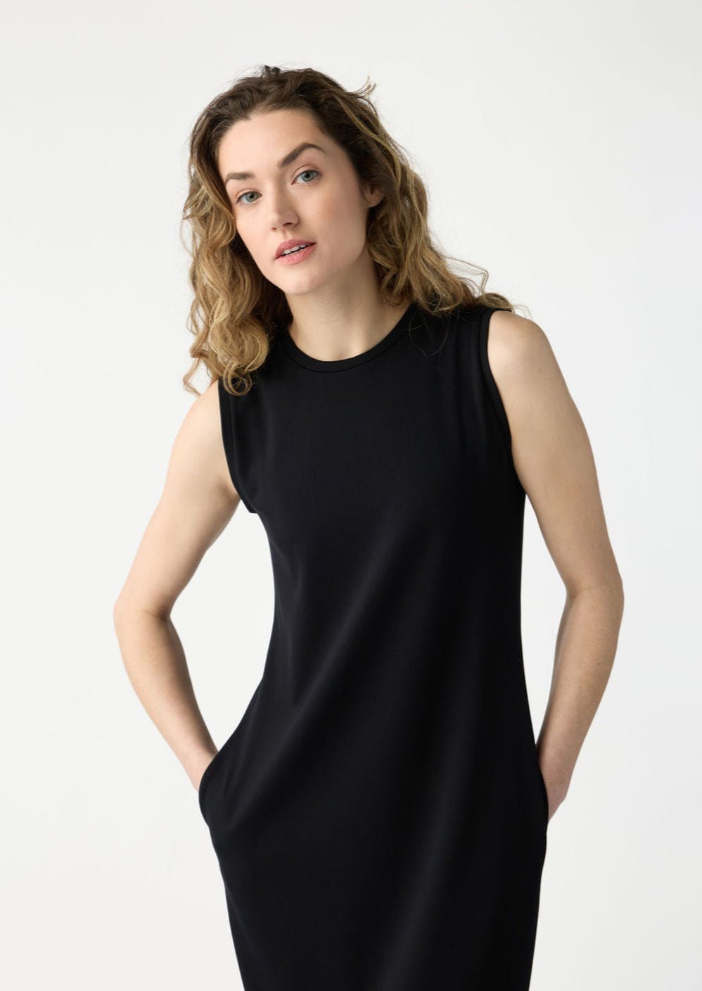 A person with long, wavy hair is wearing the Women's Brushed Bamboo Sleeveless Midi Dress by Cozy Earth in black, hands in pockets. They stand against a plain white background, looking slightly upwards with a neutral expression. |Color:Black