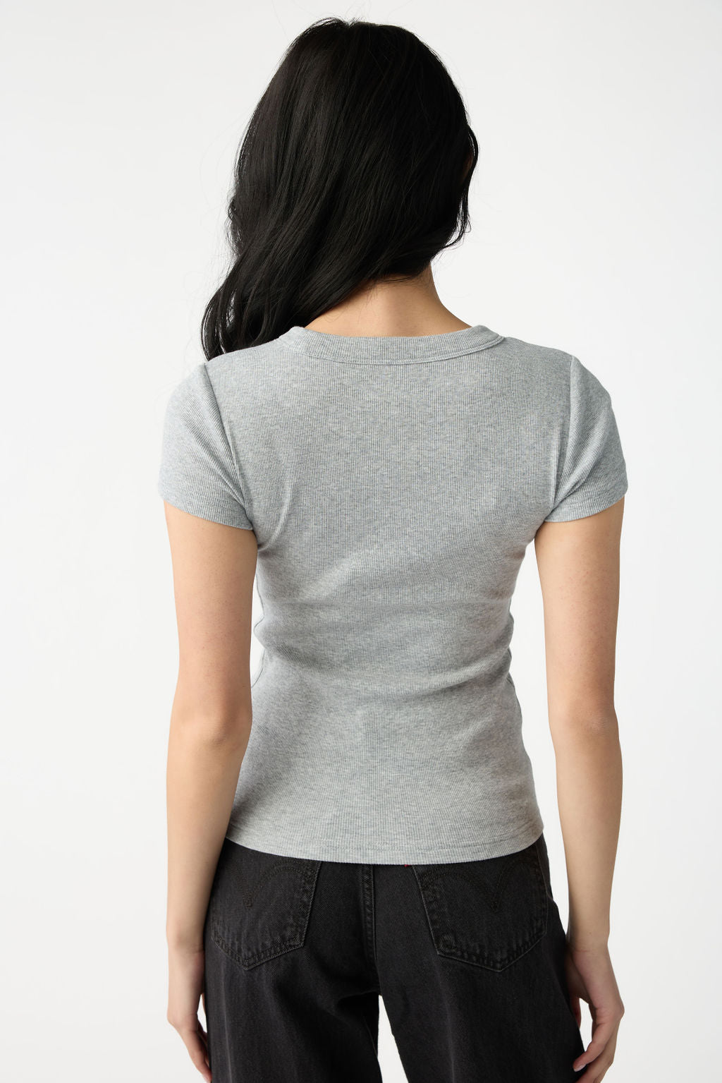 A dark haired woman wears Cozy Earth Women's Fine Ribbed Tee in Heather Grey. She is facing away from the camera and stands in front of a white background. 