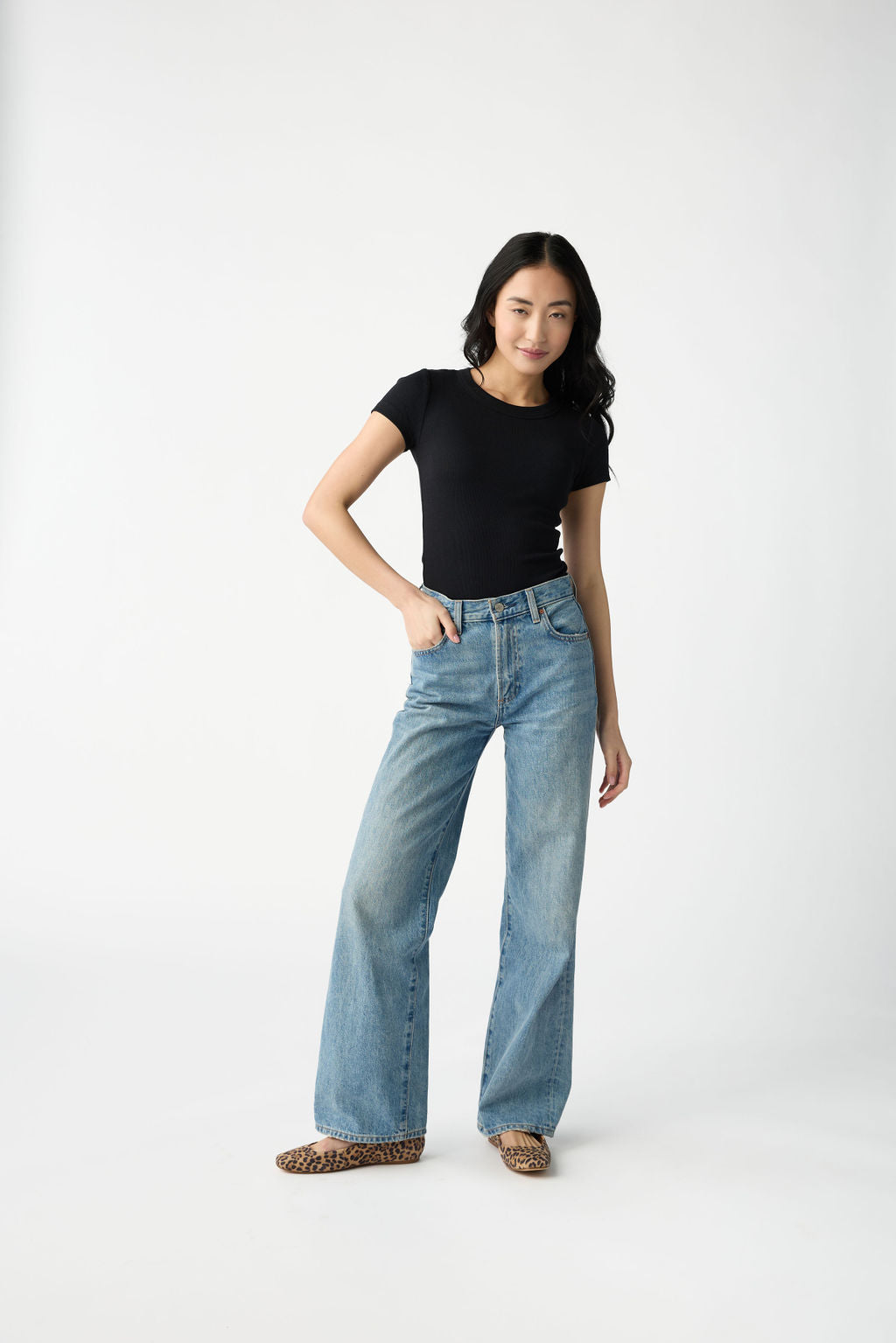 A dark haired woman wears Cozy Earth Women's Fine Ribbed Tee in Jet Black. She is wearing blue jeans and stands in front of a white background. 