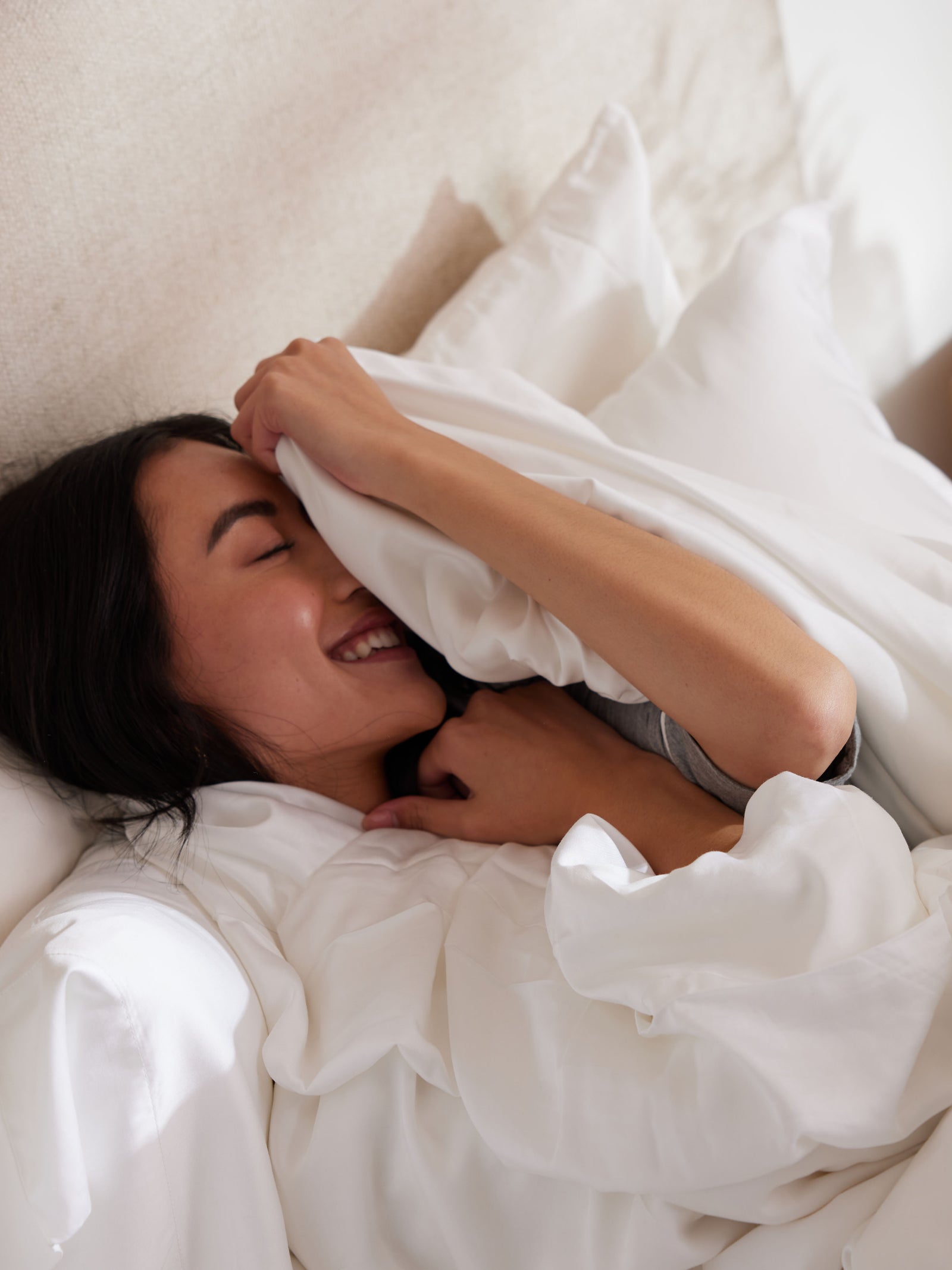 A person is lying in bed, smiling and partially covered with a Cozy Earth Silk Comforter. They are holding part of the comforter over their face, appearing relaxed and cheerful. The setting looks cozy and comfortable.