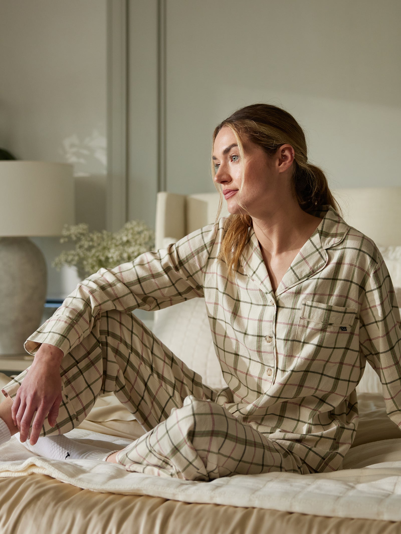 A person with long hair sits on a bed wearing the Women's Farmhouse Long Sleeve Pajama Set by Cozy Earth. They rest one hand on their knee and look pensively out of the frame. The background includes a lamp, books, and flowers on a nightstand. The room has a calm, cozy atmosphere. 