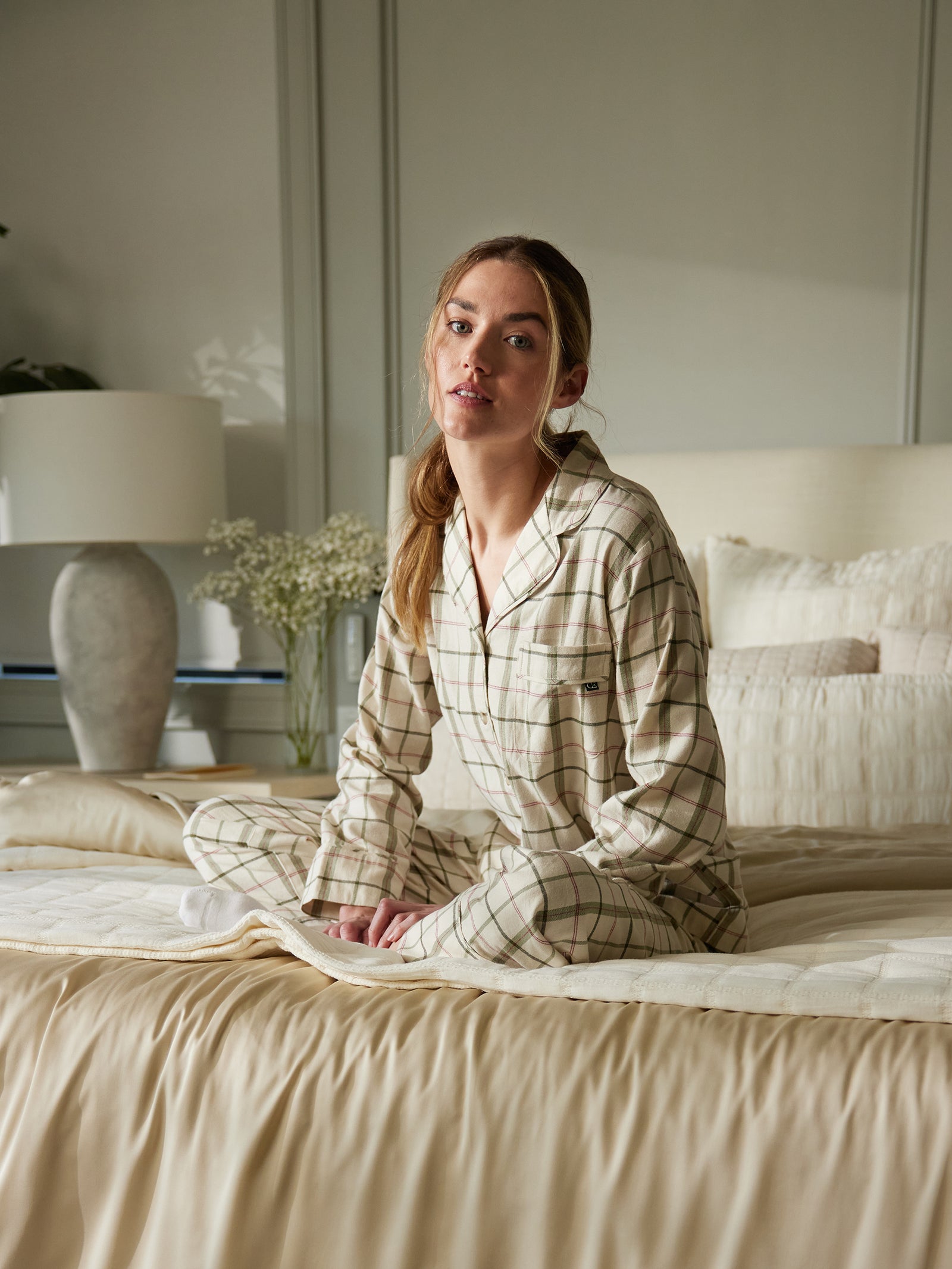 A person with long hair sits cross-legged on a bed wearing the Cozy Earth Women's Farmhouse Long Sleeve Pajama Set. The room is softly lit and includes a large vase with flowers, a table lamp, and neutral-colored bedding. Paneled walls serve as the backdrop. 