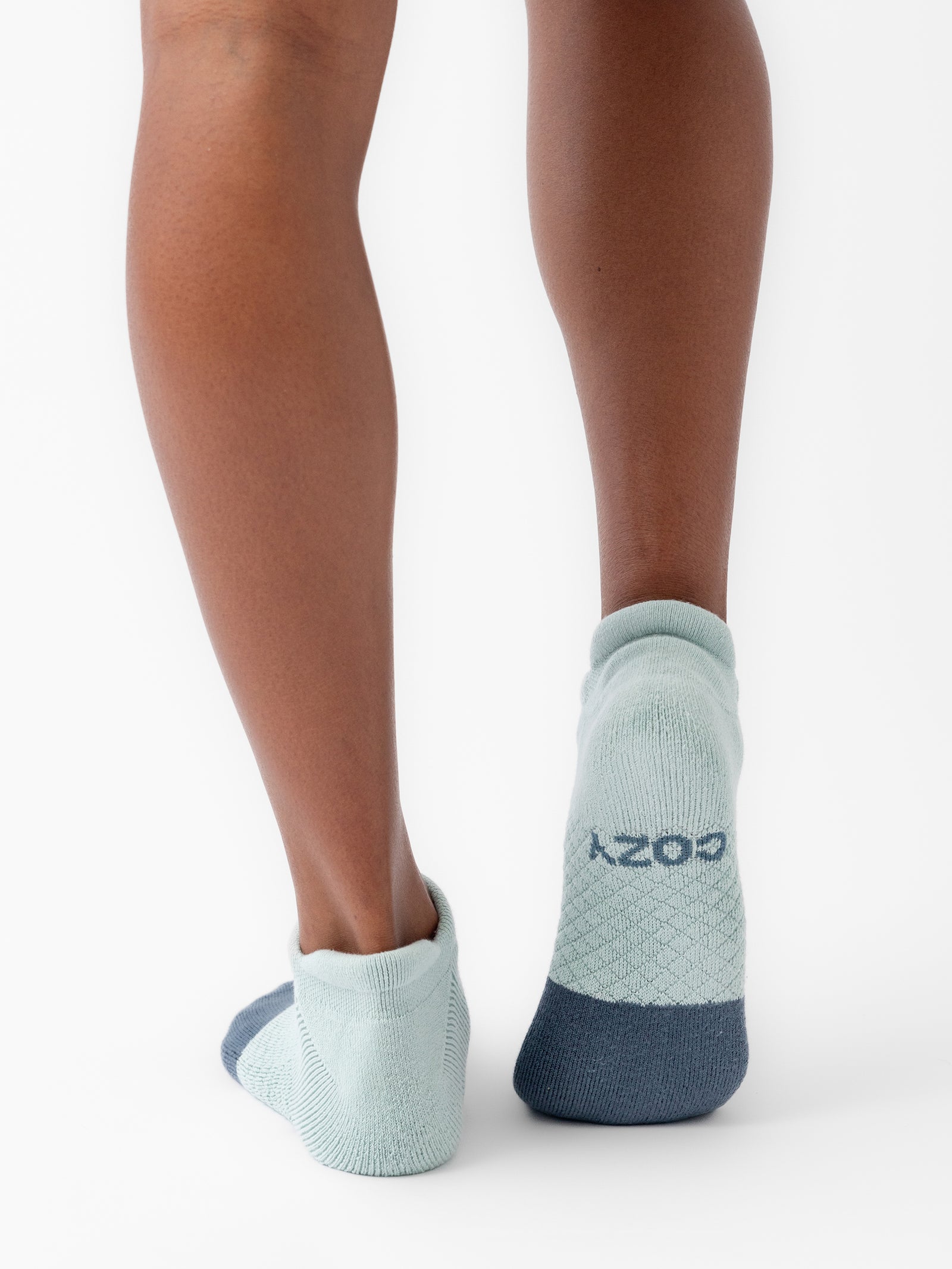 A person wearing Essential Ankle Socks by Cozy Earth, featuring a teal color with a darker teal heel and toe, stands with their back to the camera. The word "COZY" is visible on the back of one of the socks against a plain white background. 