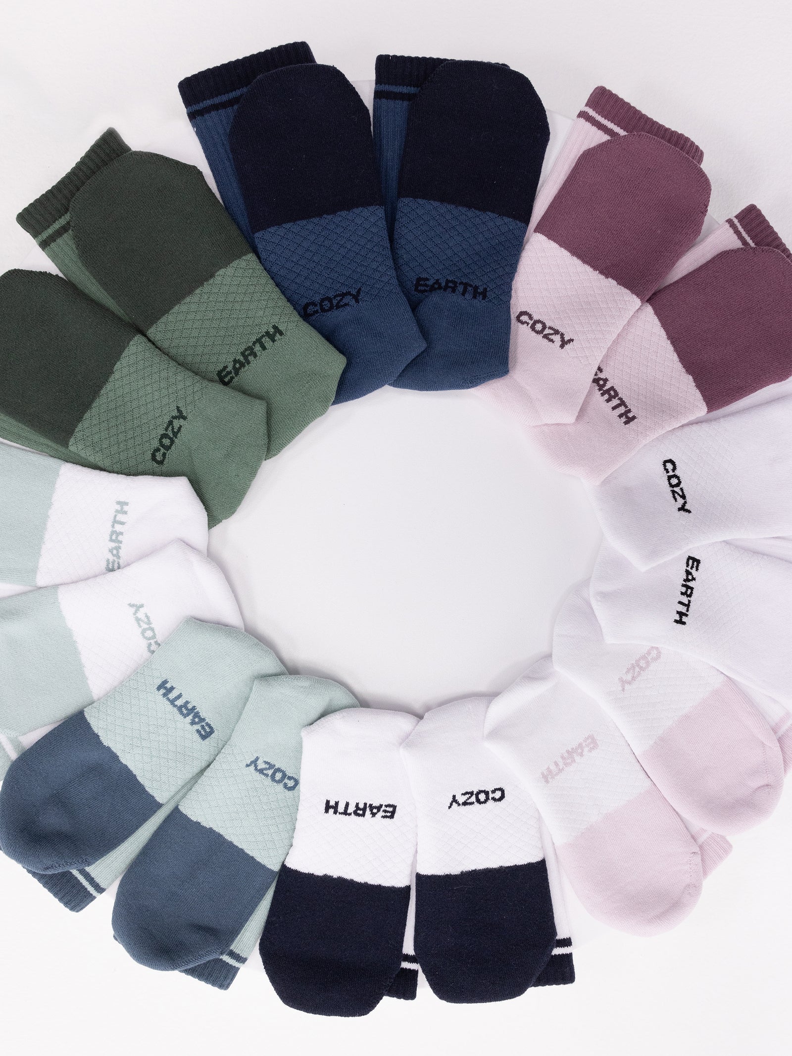 A circular arrangement of the Essential Calf Sock 2-Pack by Cozy Earth. The socks come in multiple colors, including green, navy, white, pink, and light blue. Some pairs feature text such as "EARTH" and "COZY." The background is white. 