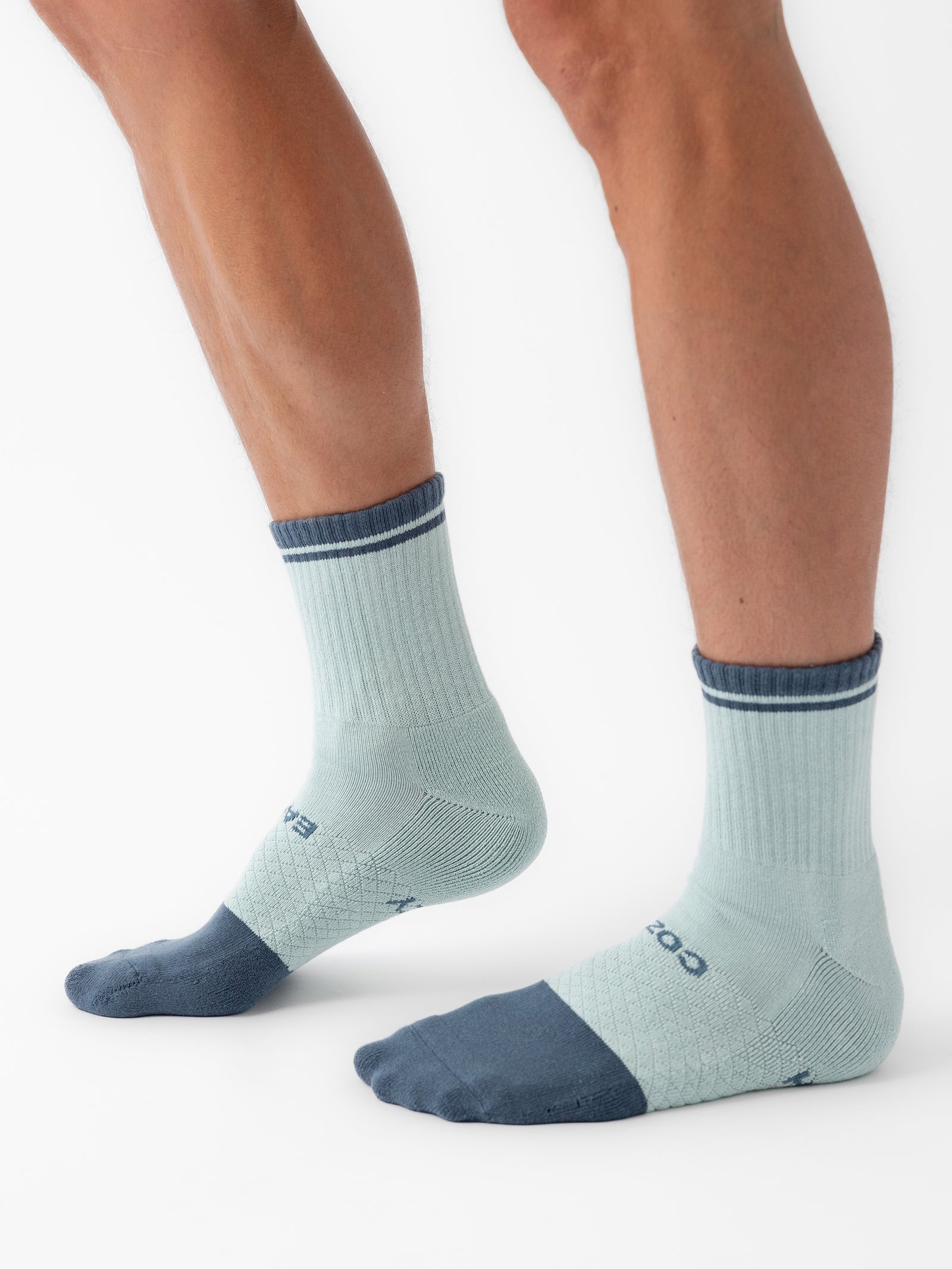 A close-up of a person's lower legs and feet wearing Cozy Earth's Essential Quarter Socks in light blue and gray, featuring a ribbed upper section and cushioned soles and toes. The person is standing with one foot slightly in front of the other on a white background. 