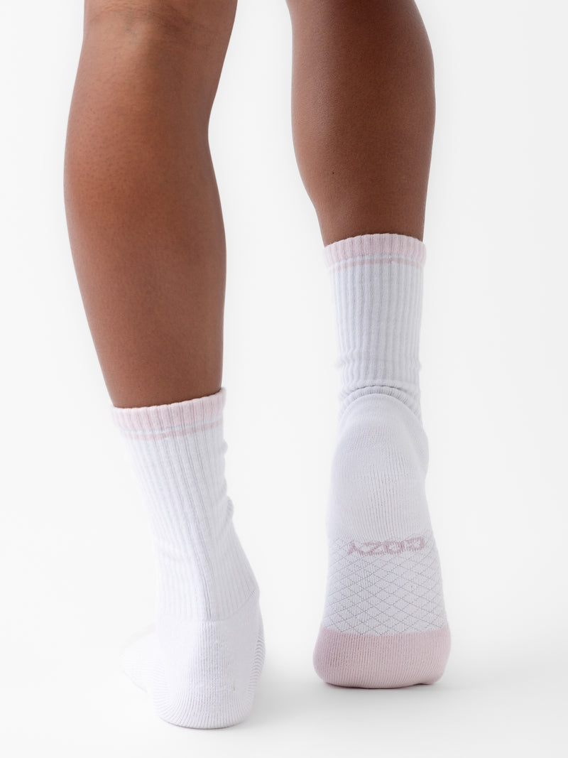 Close-up of a person's lower legs and feet wearing white Essential Calf Socks with pink accents from the Cozy Earth Essential Calf Sock 4-Pack. The left sock says "COZY" on the bottom. The plain white background emphasizes the footwear.