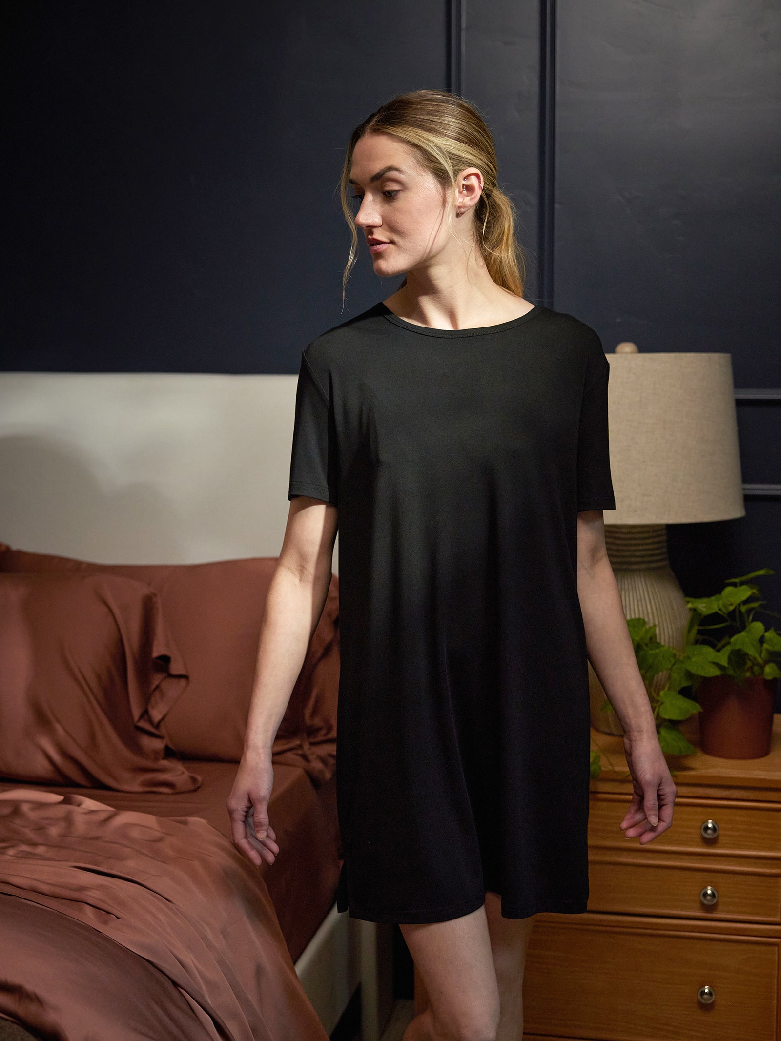 A woman wearing a Cozy Earth Women's Bamboo Stretch Knit Sleep Dress stands in a dimly lit bedroom. The bedroom features a neatly made bed with brown sheets, a wooden nightstand with a lamp, and a small plant. The walls are dark-colored, creating a cozy atmosphere. 