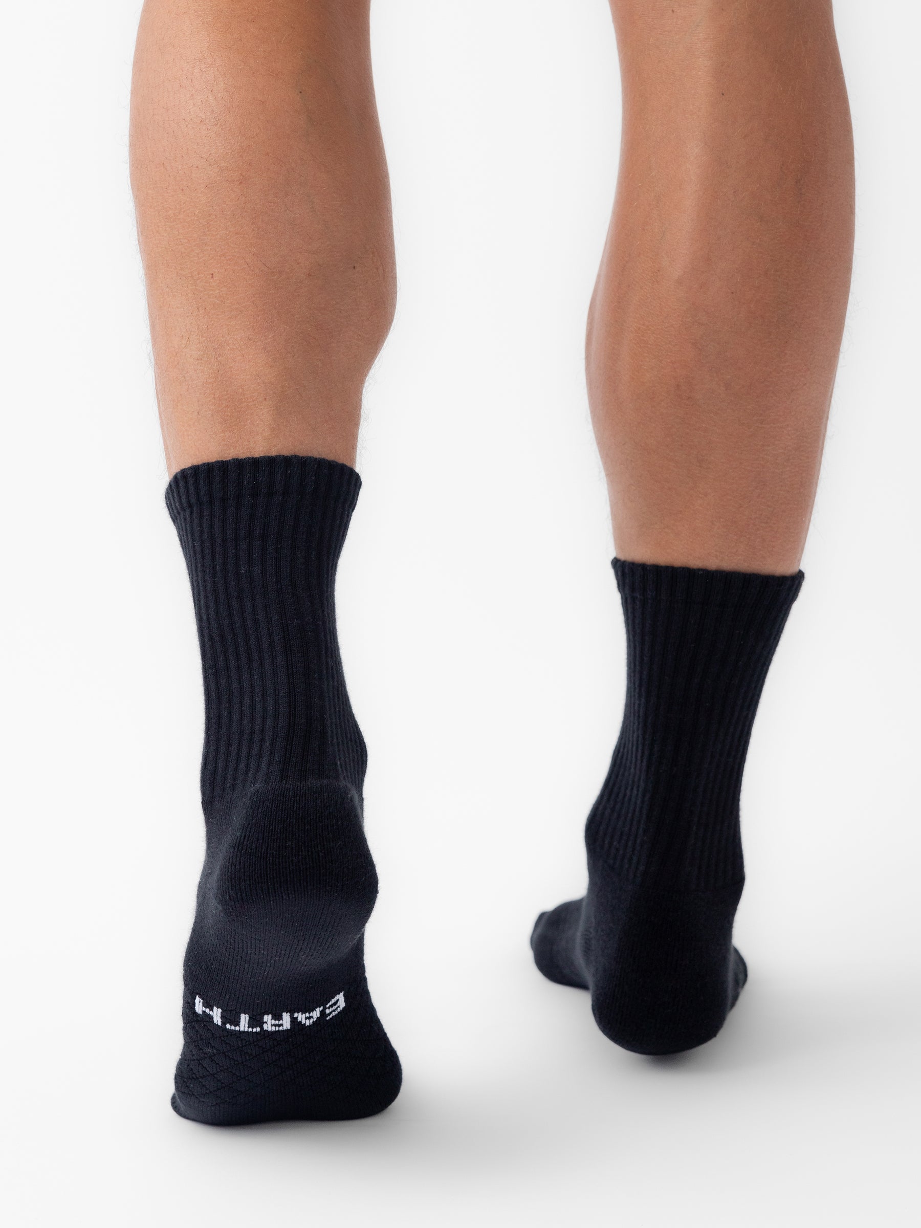 A person stands on a white surface, shown from behind, wearing black Cozy Earth Essential Calf Socks. The left sock displays "EARTH" in white letters on the sole. 