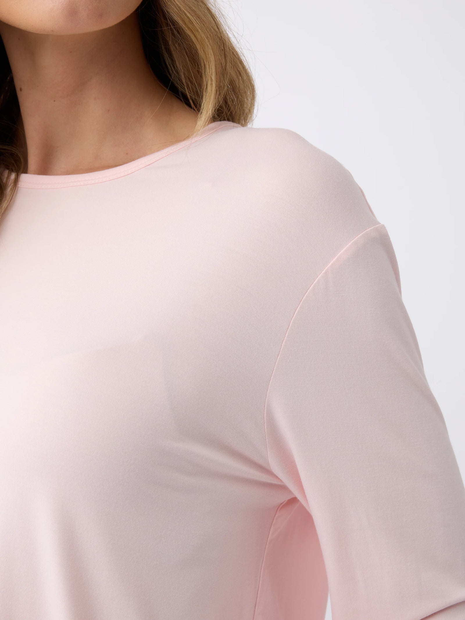 A close-up of someone in a light pink Women's Stretch Knit Bamboo Long Sleeve Lounge Tee by Cozy Earth, focusing on the upper torso and partially visible long, wavy hair. The tee features a simple round neckline. 