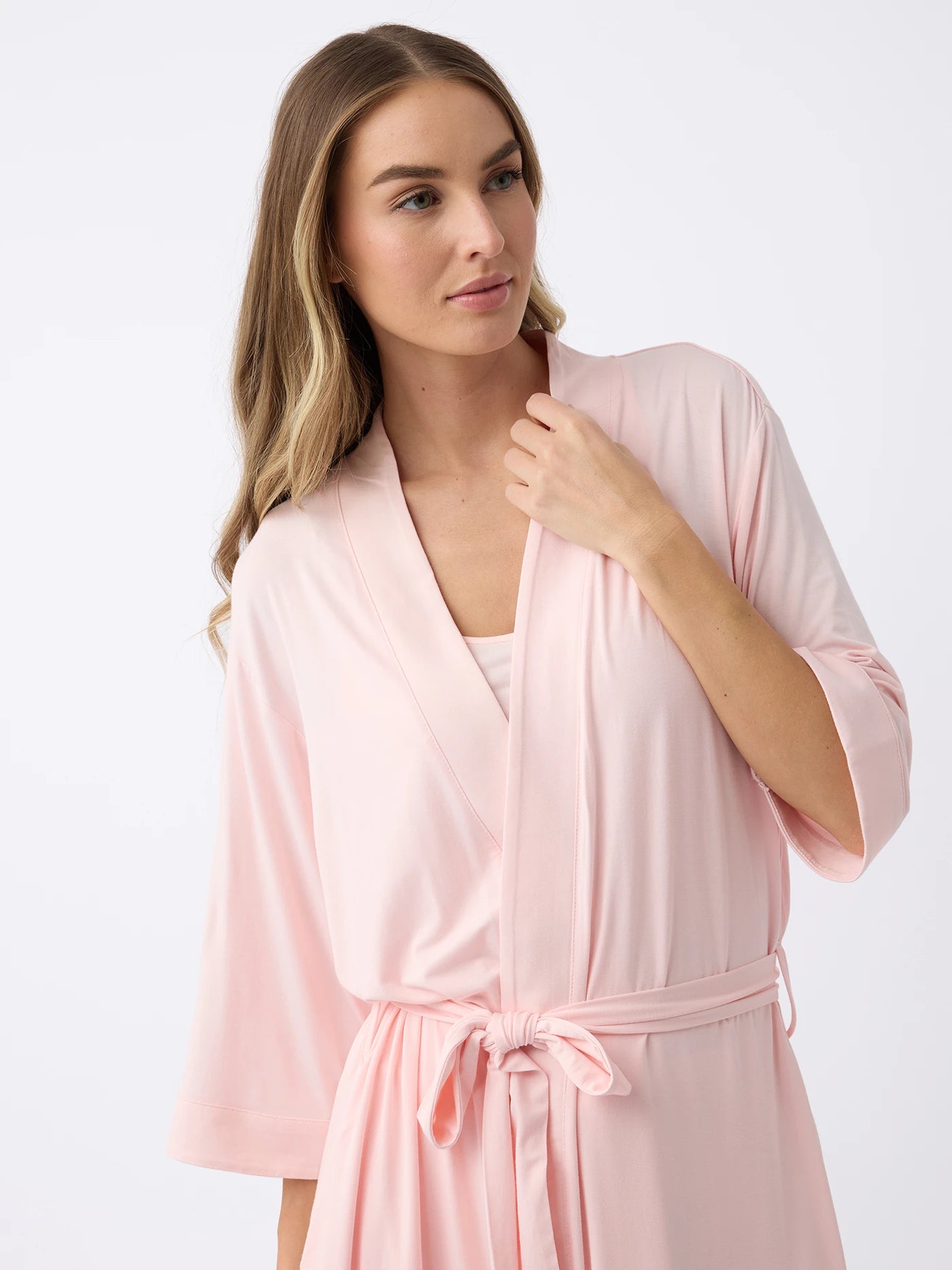 A person with long hair wears a light pink Women's Stretch-Knit Bamboo Kimono Robe by Cozy Earth, tied at the waist against a plain background, gently touching the collar. |Color:Blossom