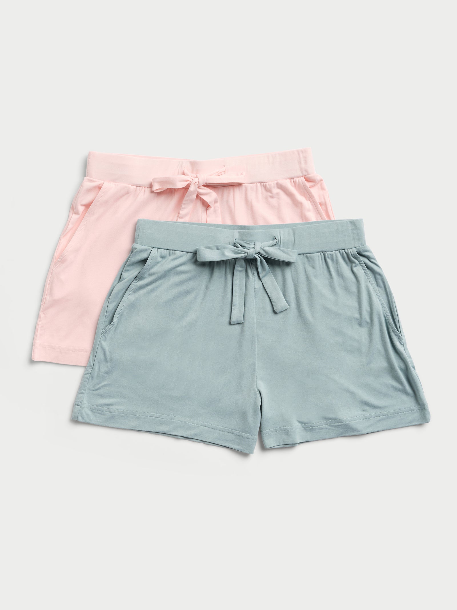 Displayed are two pairs of Cozy Earth's Stretch Knit Bamboo Shorts, one in light pink and the other in light blue. Both feature an elastic waistband with a tie front and are crafted from a soft-looking fabric. The shorts are laid flat against a light background. 