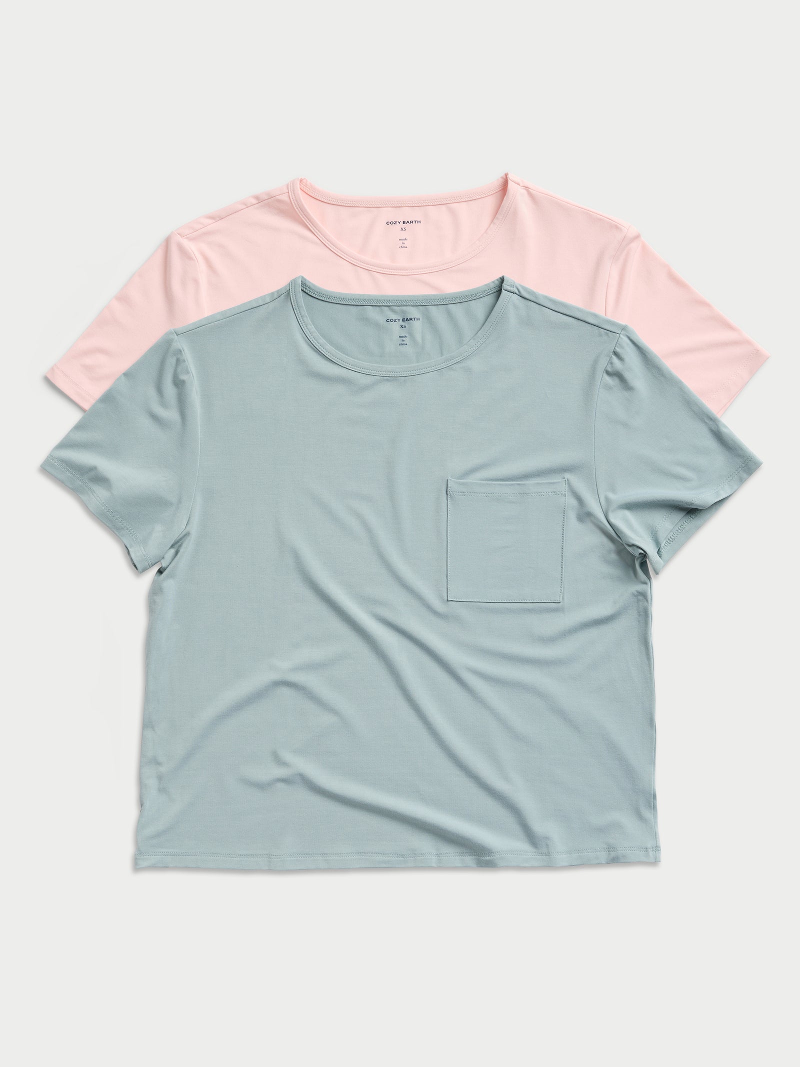 Two Women's Stretch-Knit Bamboo Lounge Tees from Cozy Earth are displayed on a flat surface. The front tee is light blue with a chest pocket, and the back tee is light pink. Both feature a round neckline and short sleeves. 