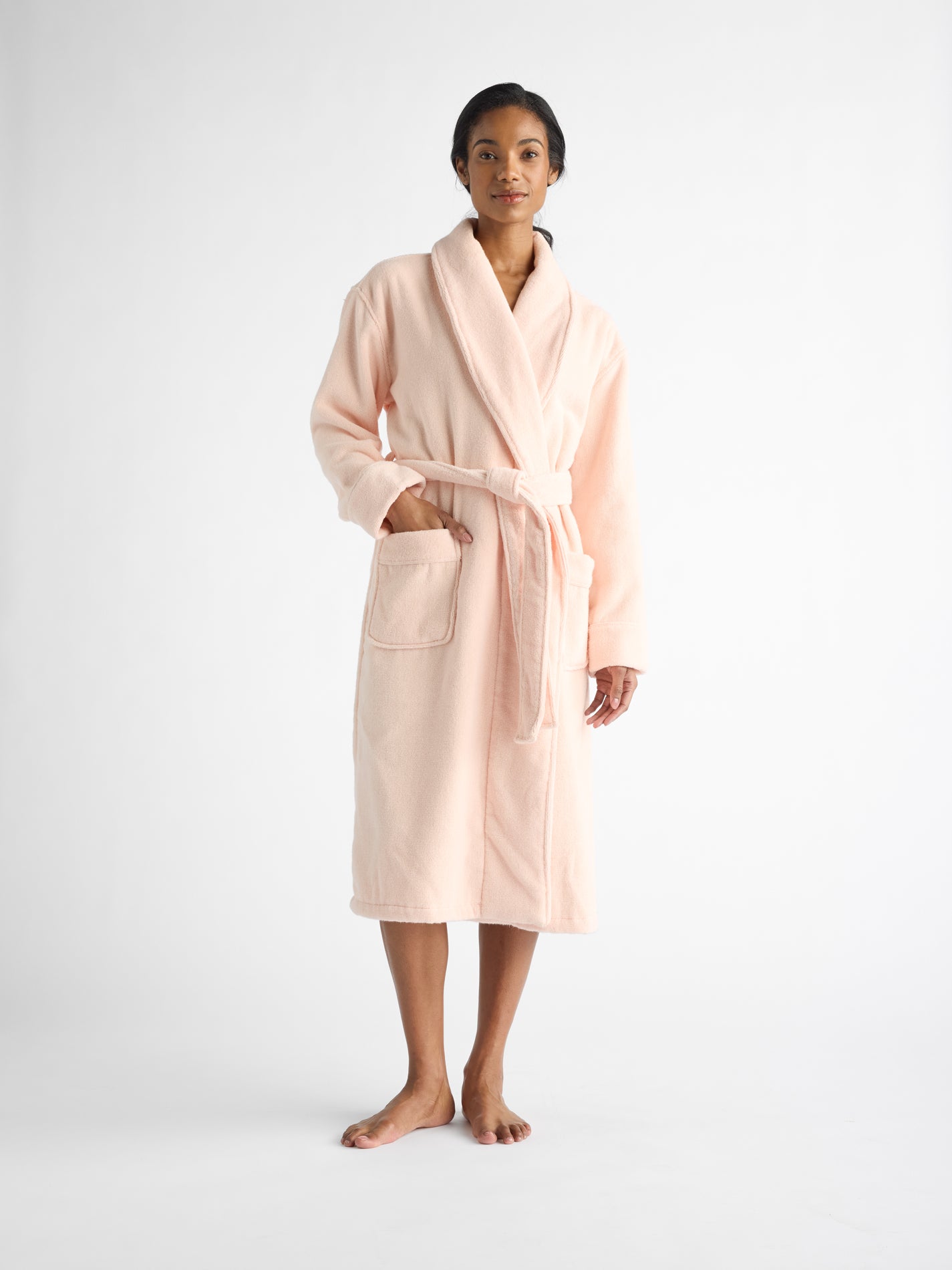 A person models the Luxe Bath Robe by Cozy Earth in soft, light pink. The robe features long sleeves, a waist tie belt, and two front pockets, set against a plain white backdrop. |Color:Blossom