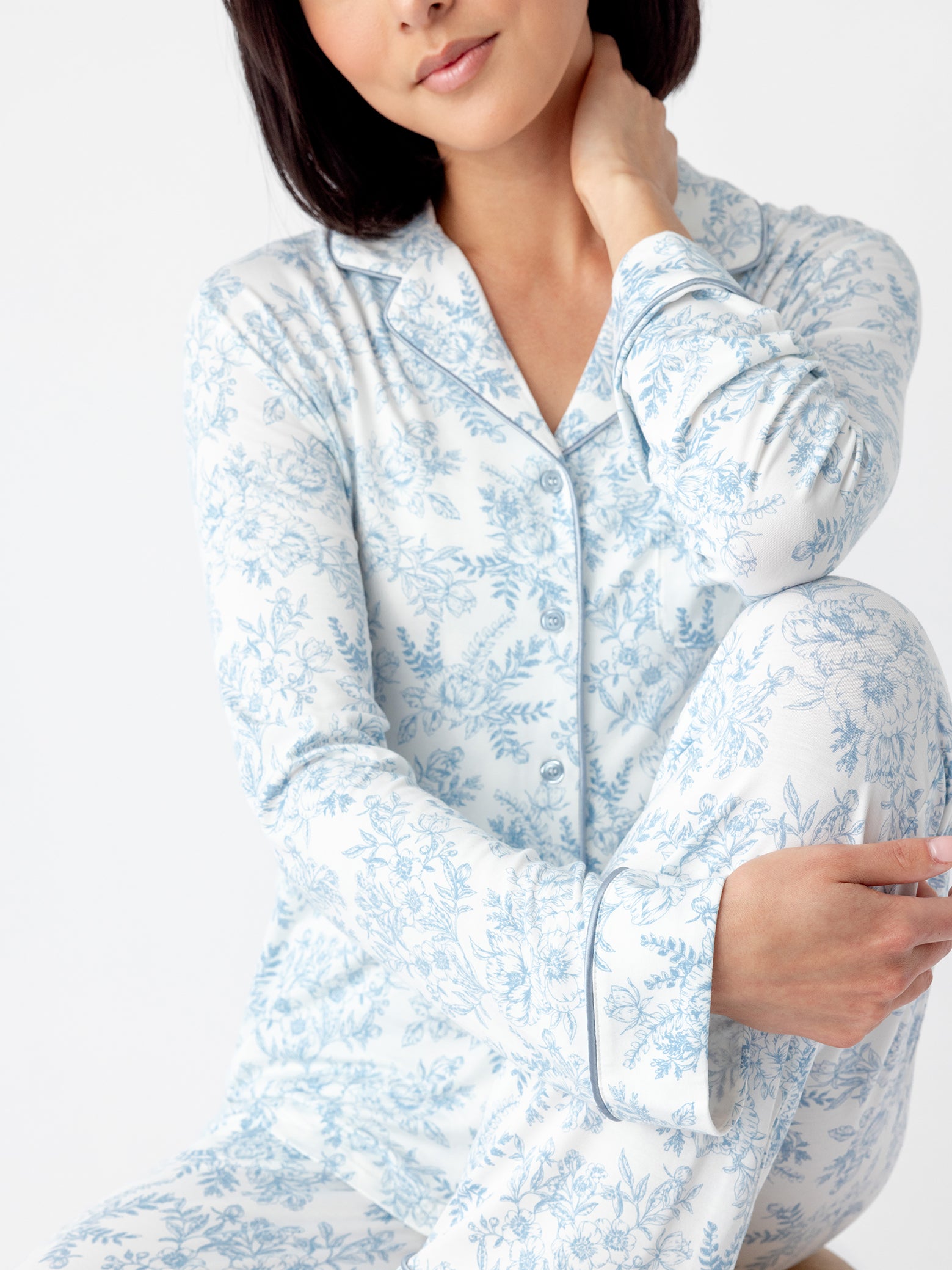 Someone wearing Cozy Earth Women's Bamboo Stretch-Knit Long Sleeve Pajama Top in a white and blue floral design sits cross-legged against a plain background. They look relaxed, with one hand resting on their head and the other on their knee.