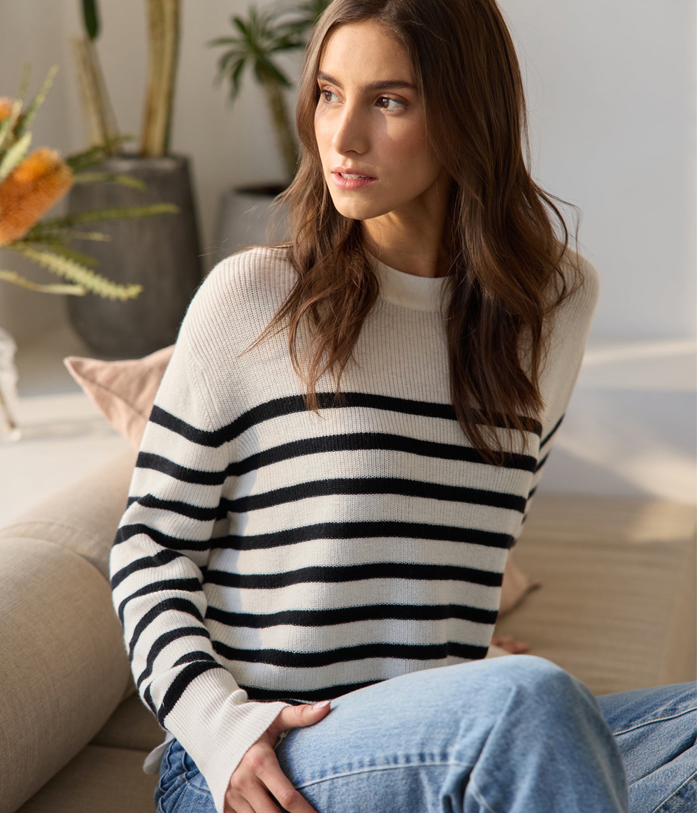 A woman with long brown hair sits on a couch, wearing a Cozy Earth Women's Rowan Sweater and blue jeans. She gazes sideways, with a potted plant and soft natural light in the background. 