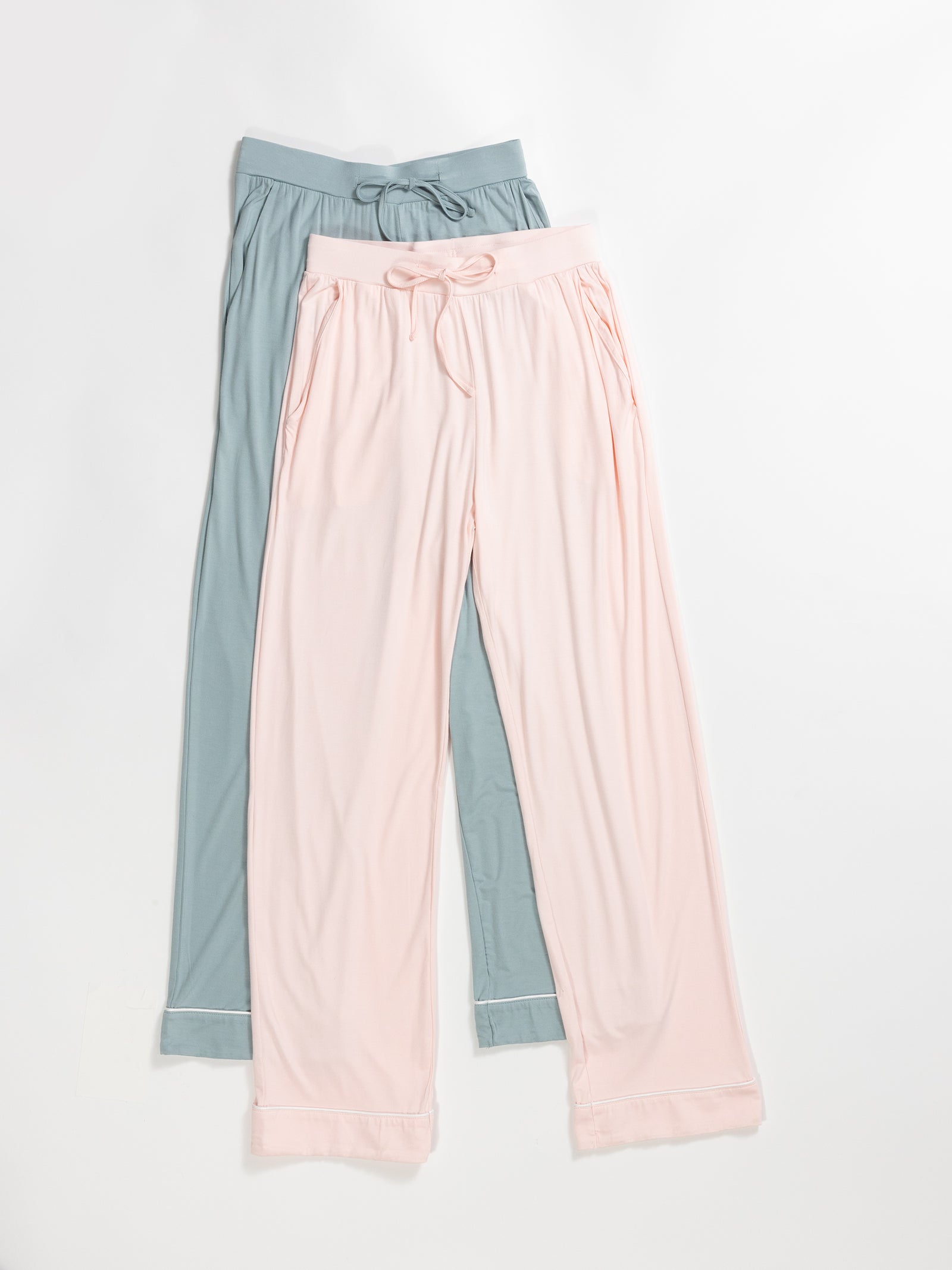 Two pairs of Women's Stretch-Knit Bamboo Pajama Pants by HIDE are laid flat on a light surface. One pair is a soft pink, featuring side pockets and a drawstring waist. The other pair is a muted teal color, also with pockets and a drawstring waist. Both have a relaxed, comfortable fit. 