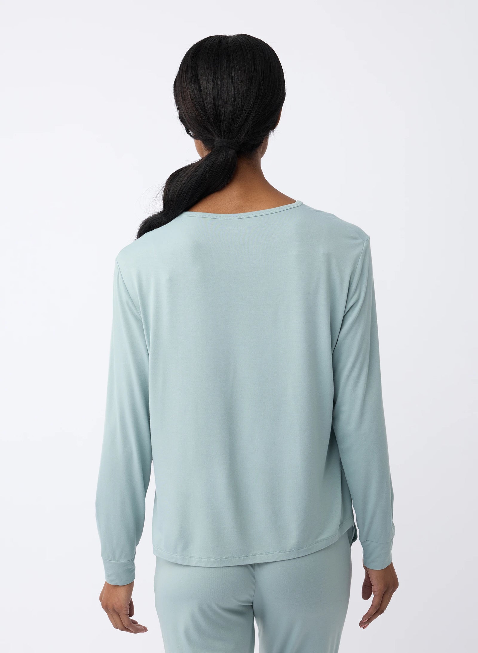 A woman with long dark hair, seen from behind, is wearing a Cozy Earth Women's Stretch Knit Bamboo Long Sleeve Lounge Tee in light blue and matching pants against a plain white background. 