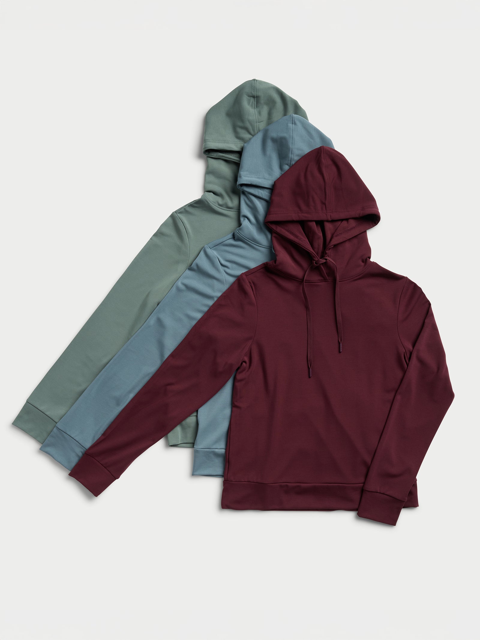 A stack of four Women's Ultra-Soft Bamboo Hoodies by Cozy Earth is neatly displayed on a plain background, showcasing the soft fabric and casual style in muted shades of green, blue, and maroon with their hoods prominently visible. 