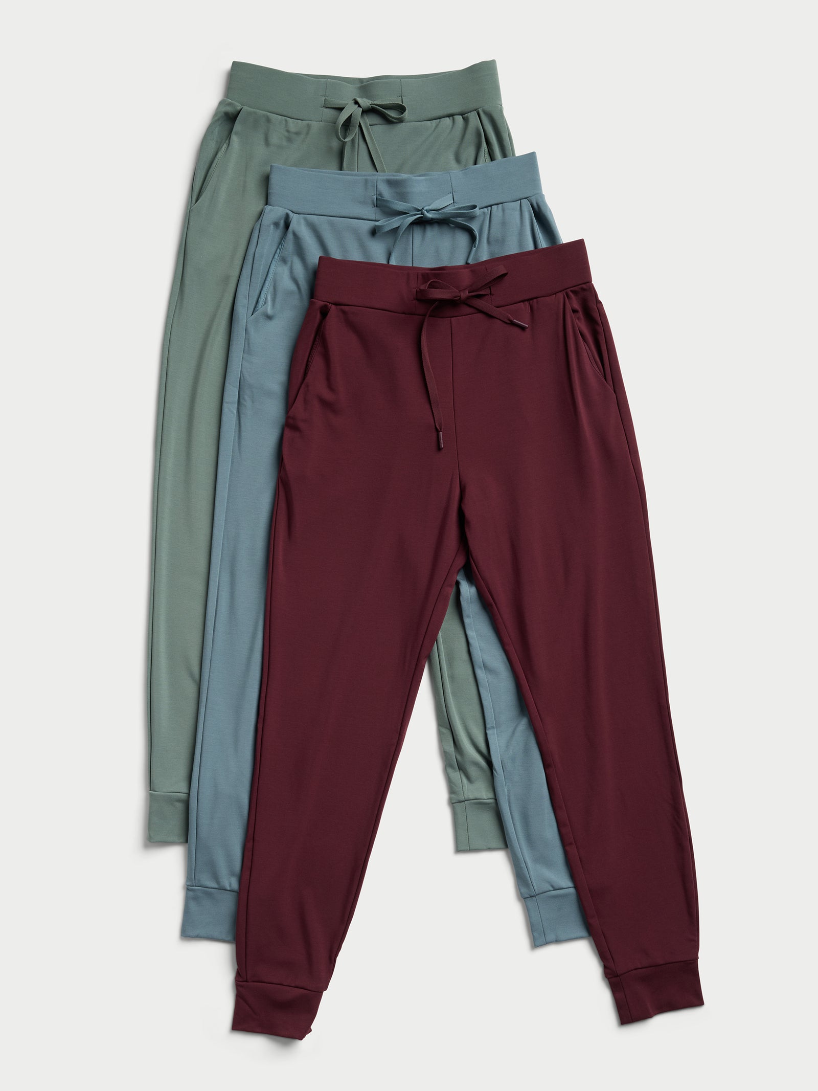 Three pairs of Women's Bamboo Jogger Pants by Cozy Earth in green, teal, and burgundy are laid flat on a light background. Each pair showcases drawstrings and elastic waistbands, complemented by ribbed cuffs at the ankles. The pants are neatly overlapped in a vertical arrangement. 