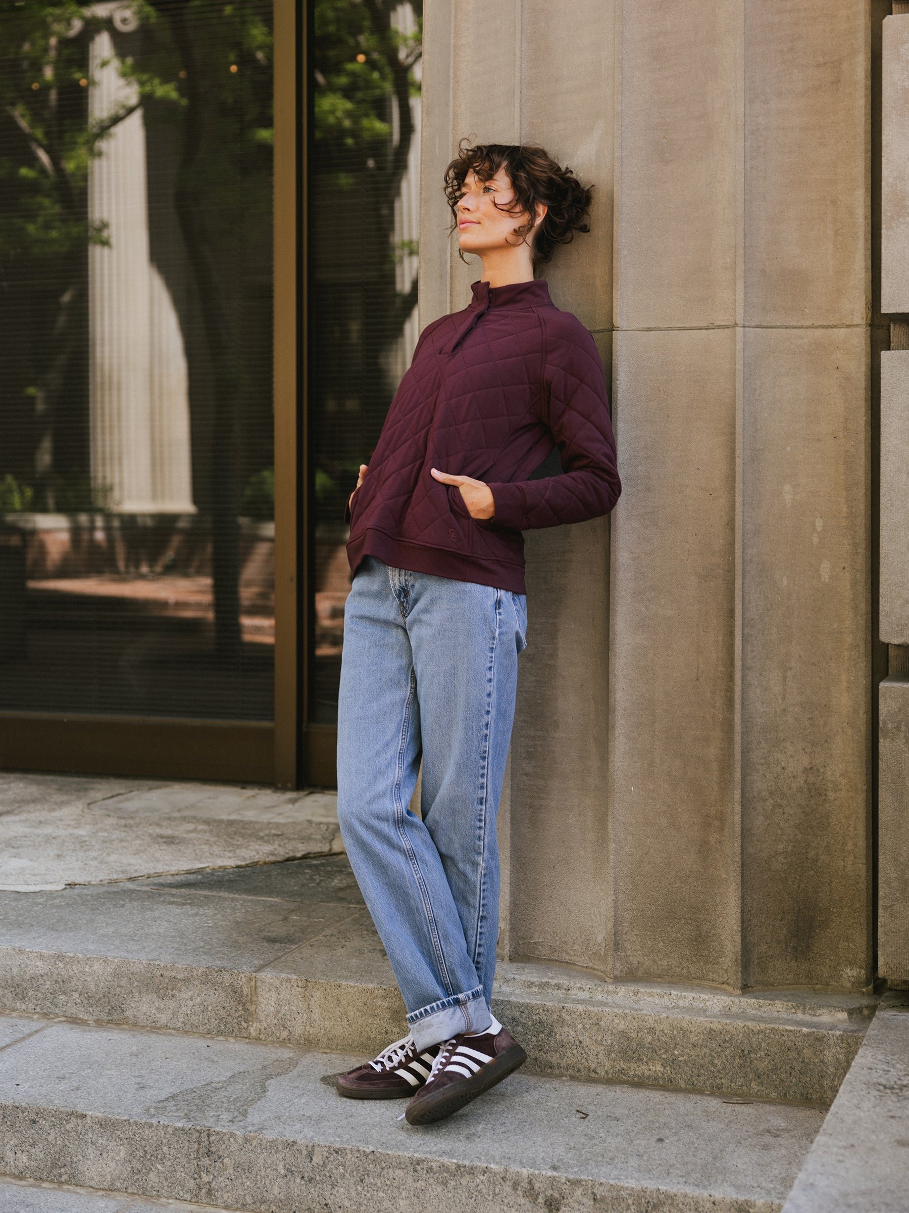 A person with short curly hair stands against a stone wall outdoors, wearing a maroon Women's Ultra-Soft Bamboo Quilted Snap Pullover from Cozy Earth, light blue jeans, and maroon sneakers with white stripes. They have their hands in their jacket pockets and gaze upwards with a relaxed expression. 