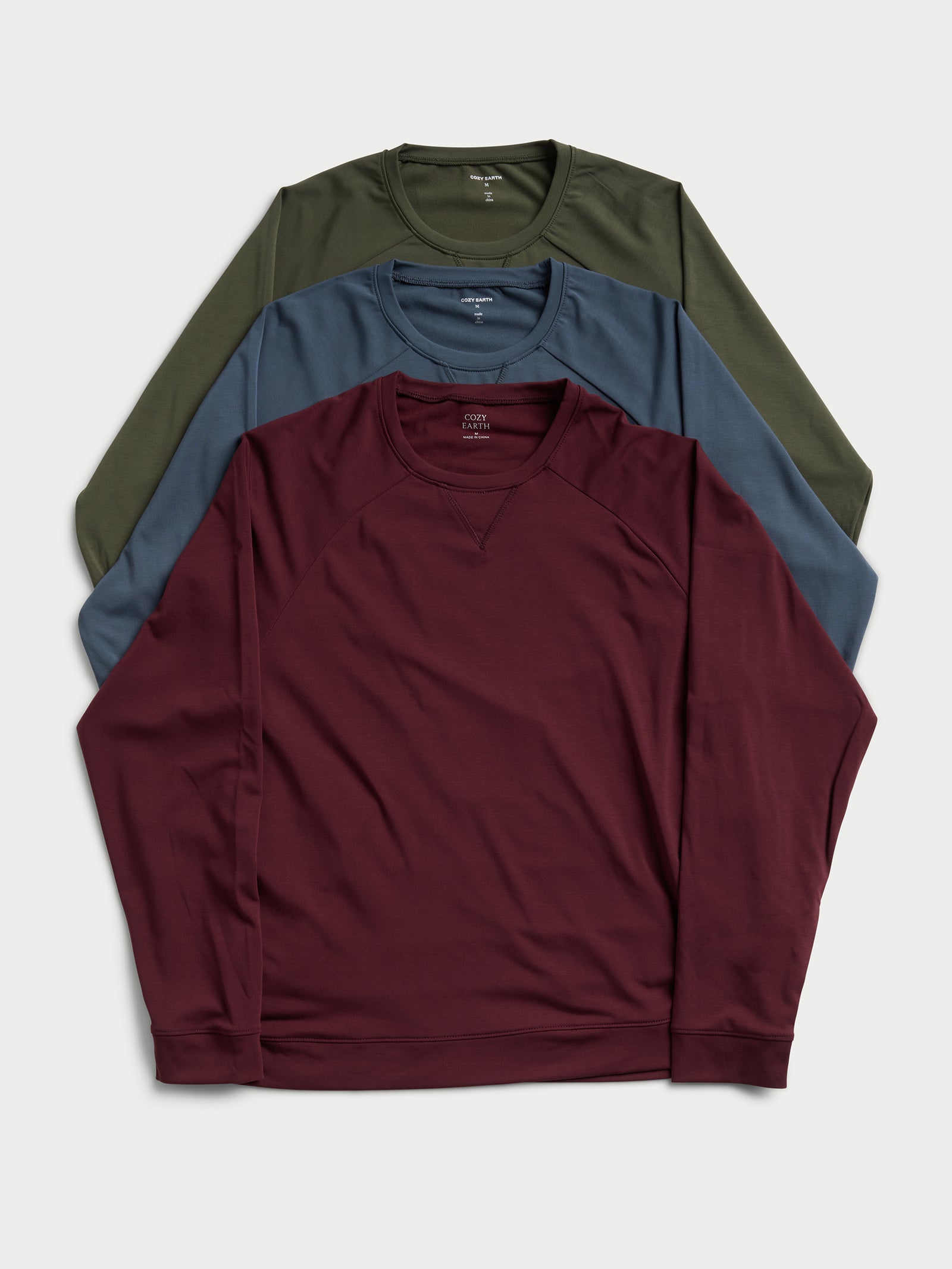 Three Men's Ultra-Soft Bamboo Pullover Crews by Cozy Earth are arranged in a layered fashion: the dark red one is at the front, the blue one is positioned in the middle, and the olive green one is at the back, all set against a light gray background. 