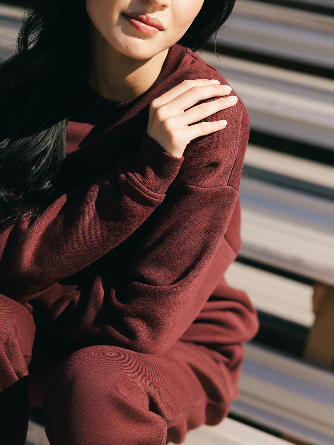 Wearing a Cozy Earth Women's CityScape Crewneck, a person is seated outdoors on wooden steps. They are touching their left shoulder with their right hand, and only the lower part of their face is visible. The lighting creates soft shadows on the ensemble. 