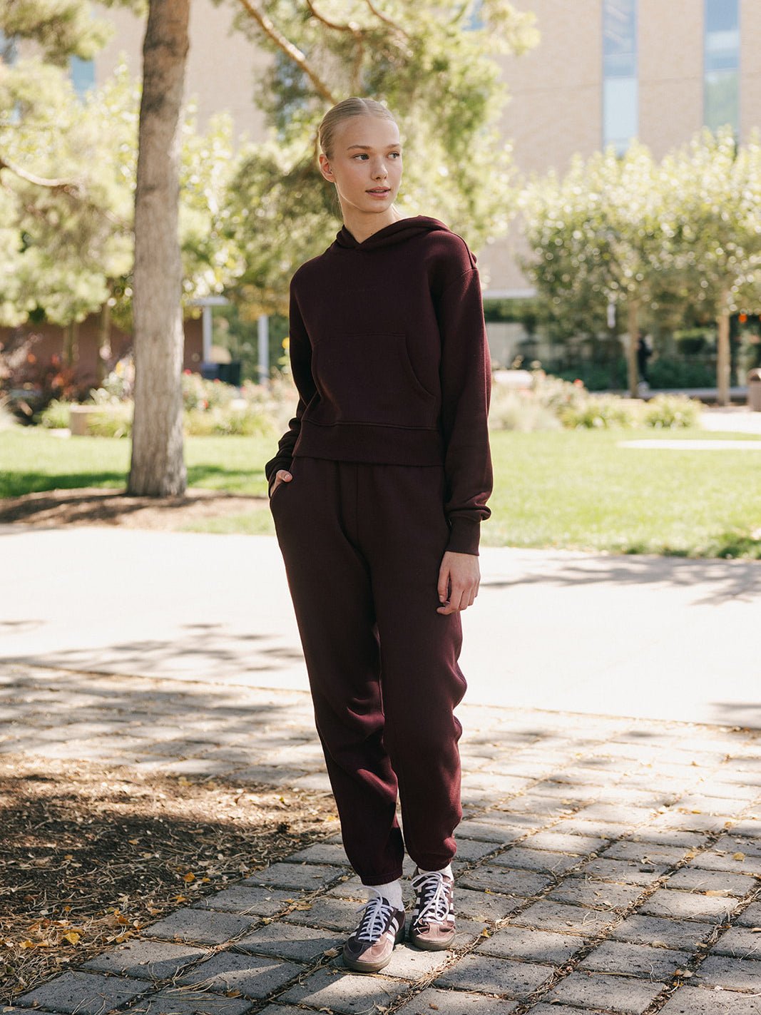 A person with light hair is standing outdoors on a paved pathway surrounded by trees and greenery. They are wearing a matching dark burgundy set, including the Women's CityScape Cropped Hoodie from Cozy Earth, and looking to the side with a relaxed expression. The background includes a building and sunlight filtering through the leaves. 