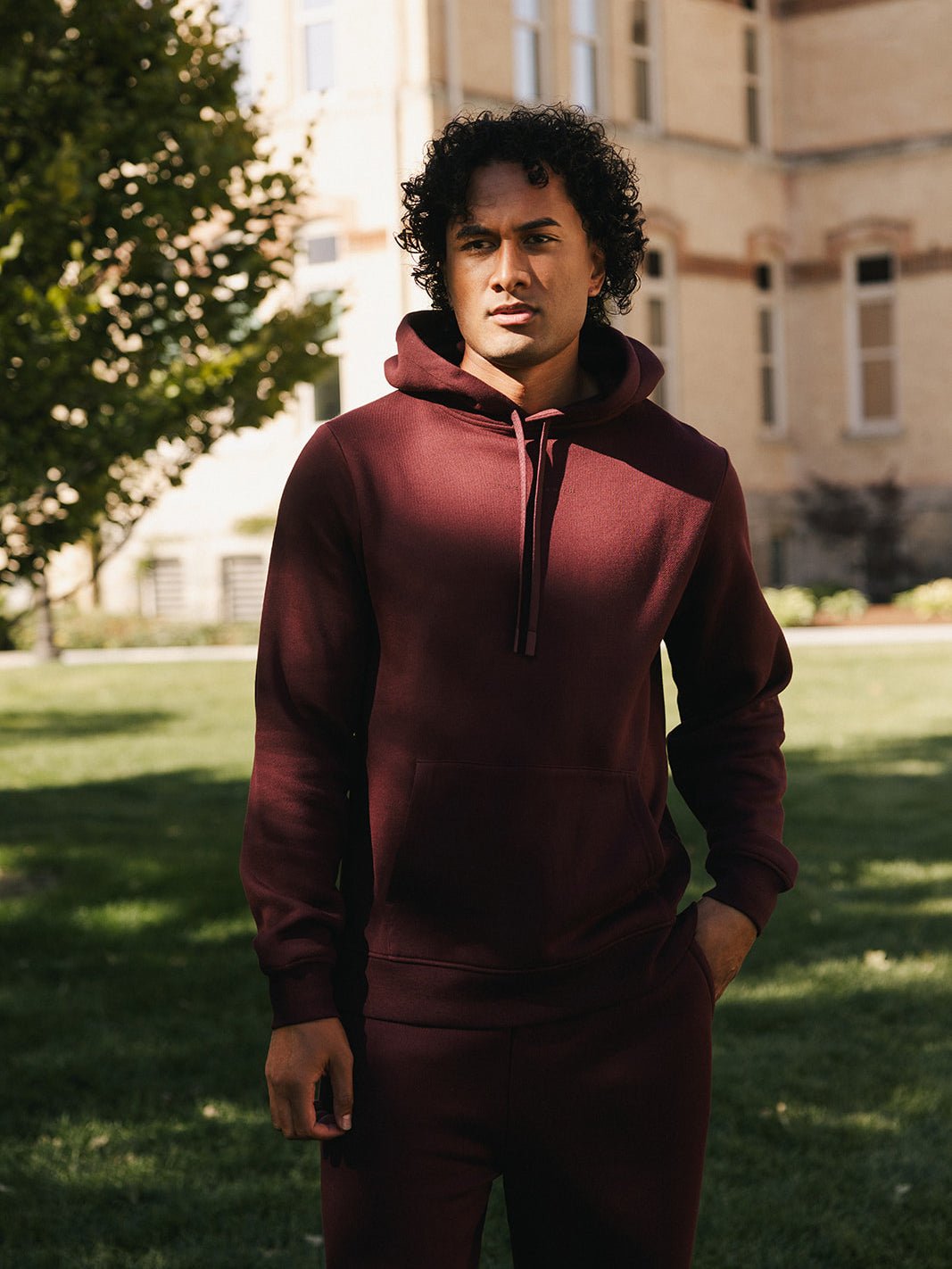 A person with curly hair wears the Cozy Earth Men's CityScape Hoodie in burgundy along with matching sweatpants, standing outside in a sunlit area surrounded by grass and trees. In the background, there's a tall building with multiple windows. They appear relaxed with one hand in their pocket. 